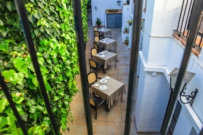 An outdoor patio with tables and chairs, surrounded by a wall of green foliage.