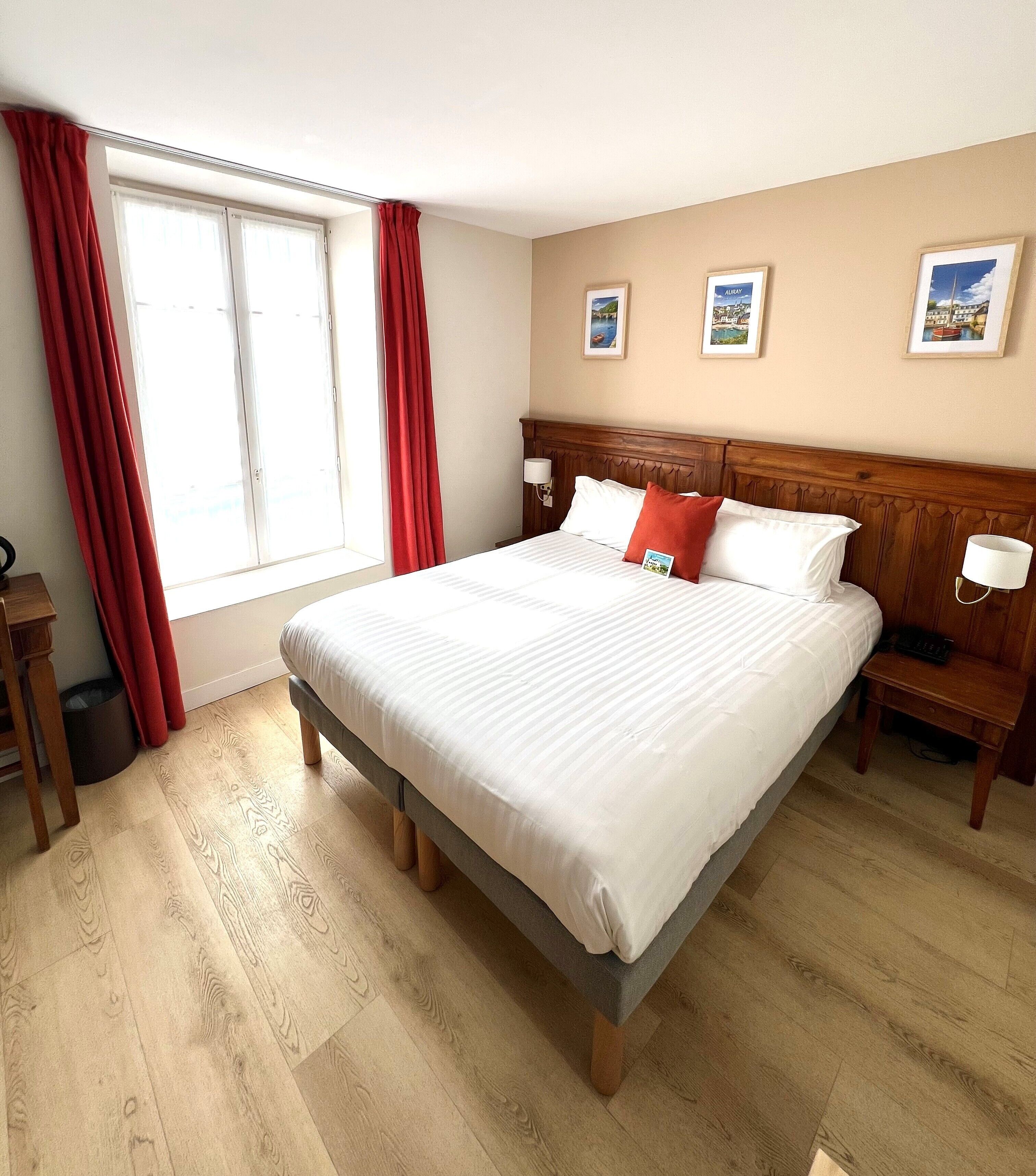 Une chambre d’hôtel avec un grand lit, une tête de lit en bois, deux tables de chevet avec des lampes, une fenêtre avec des rideaux rouges et des tableaux encadrés accrochés au mur.
