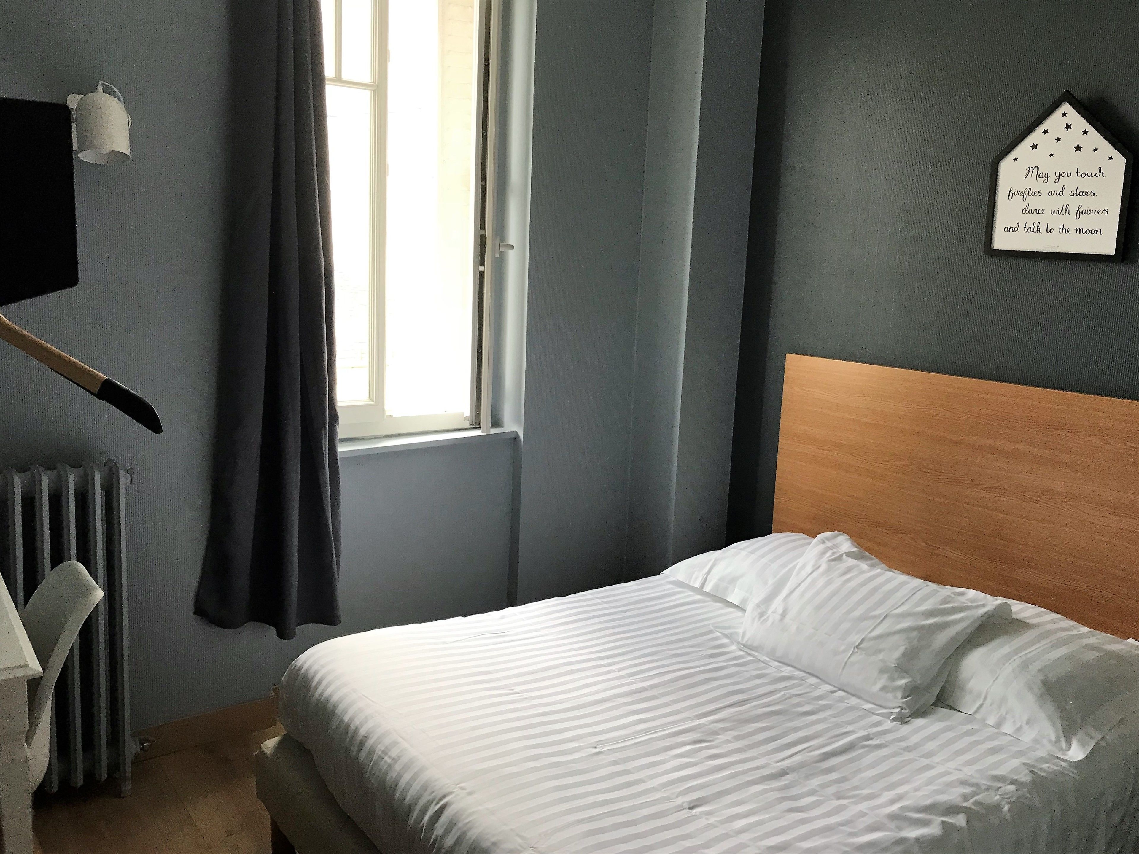 A bedroom with a bed, a window with curtains, a wall-mounted light, and a decorative sign.