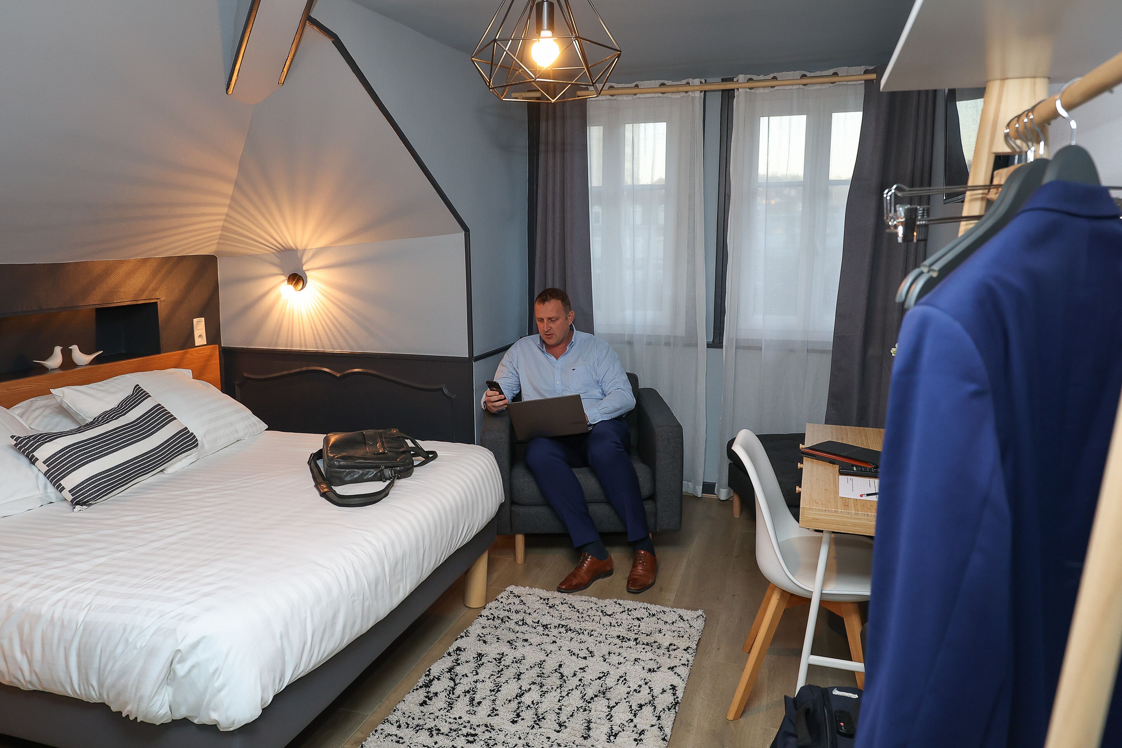 Habitación de hotel con cama, escritorio, silla, perchero y ventana con cortinas.