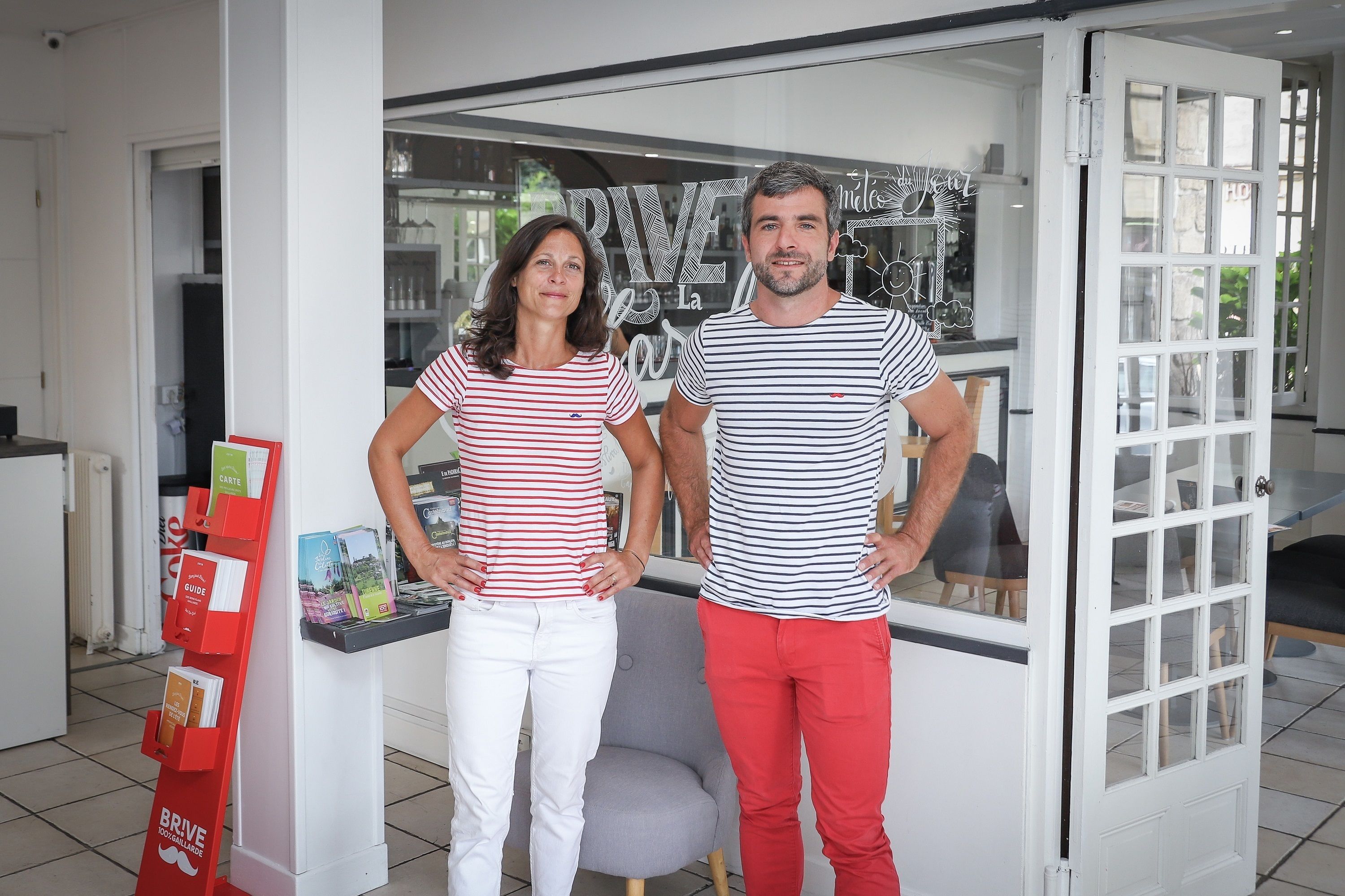 A man and a woman standing in a room with a glass door, a red display stand with brochures, and a grey sofa.
