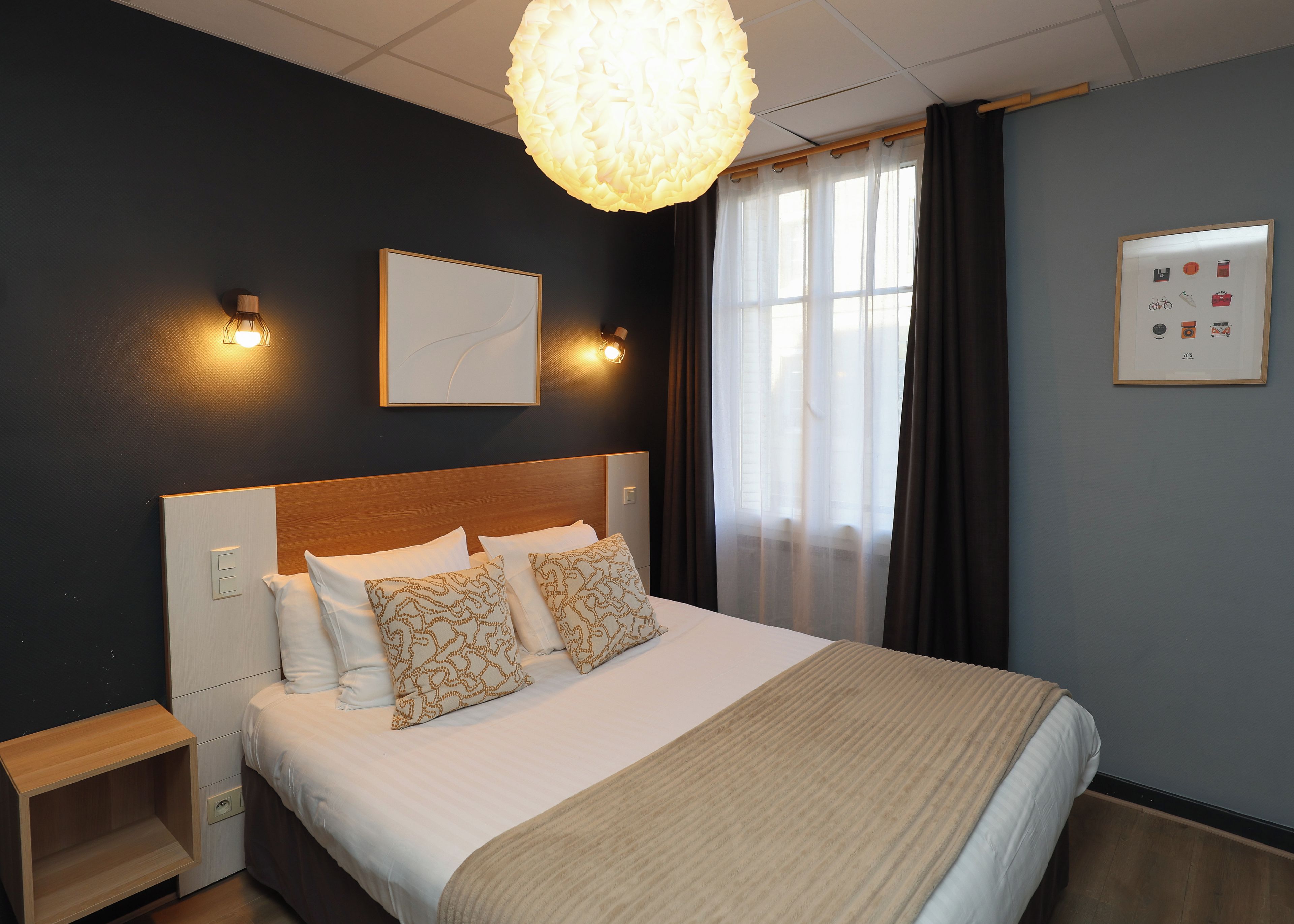 A hotel room with a bed, bedside table, wall-mounted light, and a window with curtains.