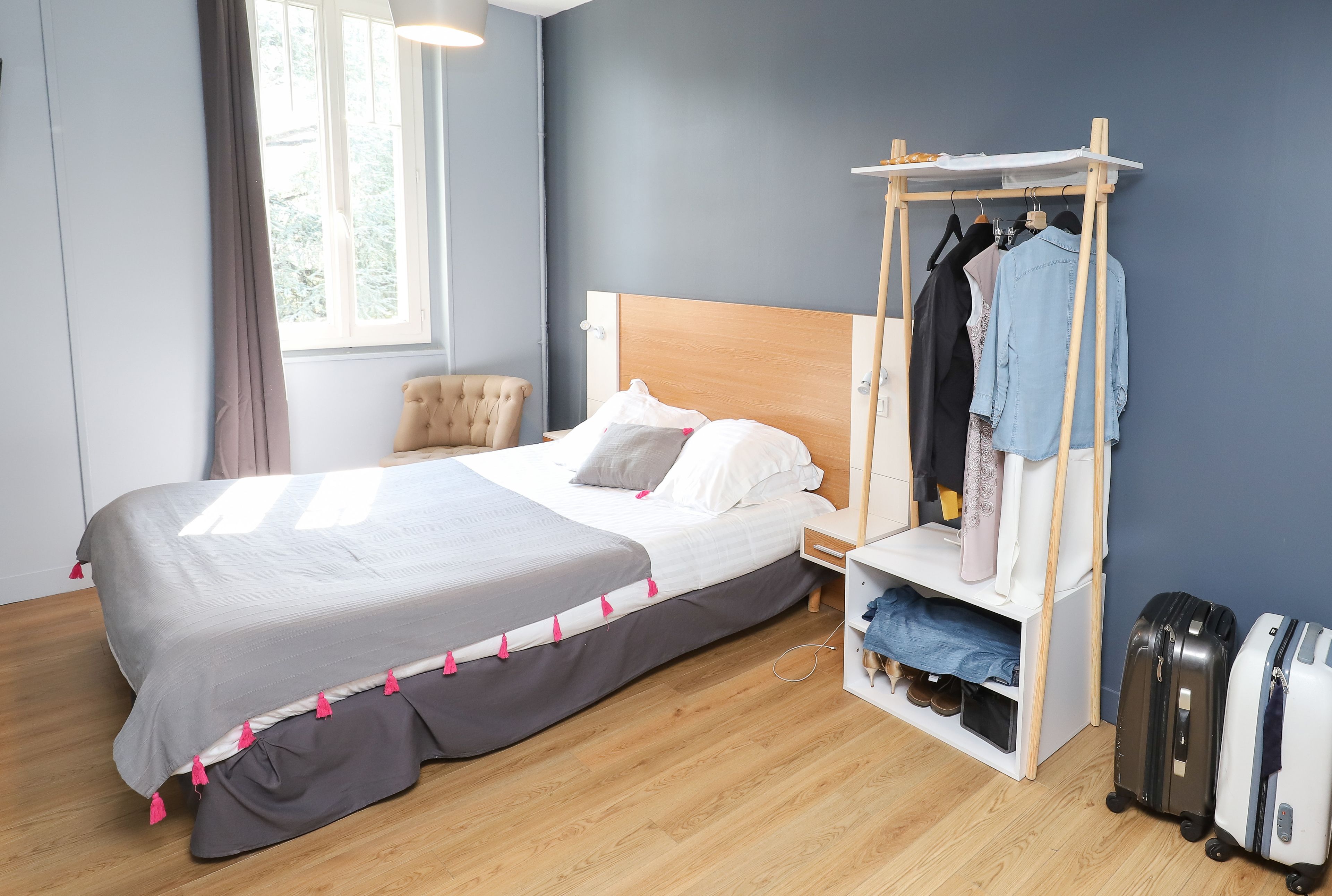 A neatly arranged hotel room with a bed, a wooden nightstand, a clothing rack, and two suitcases.