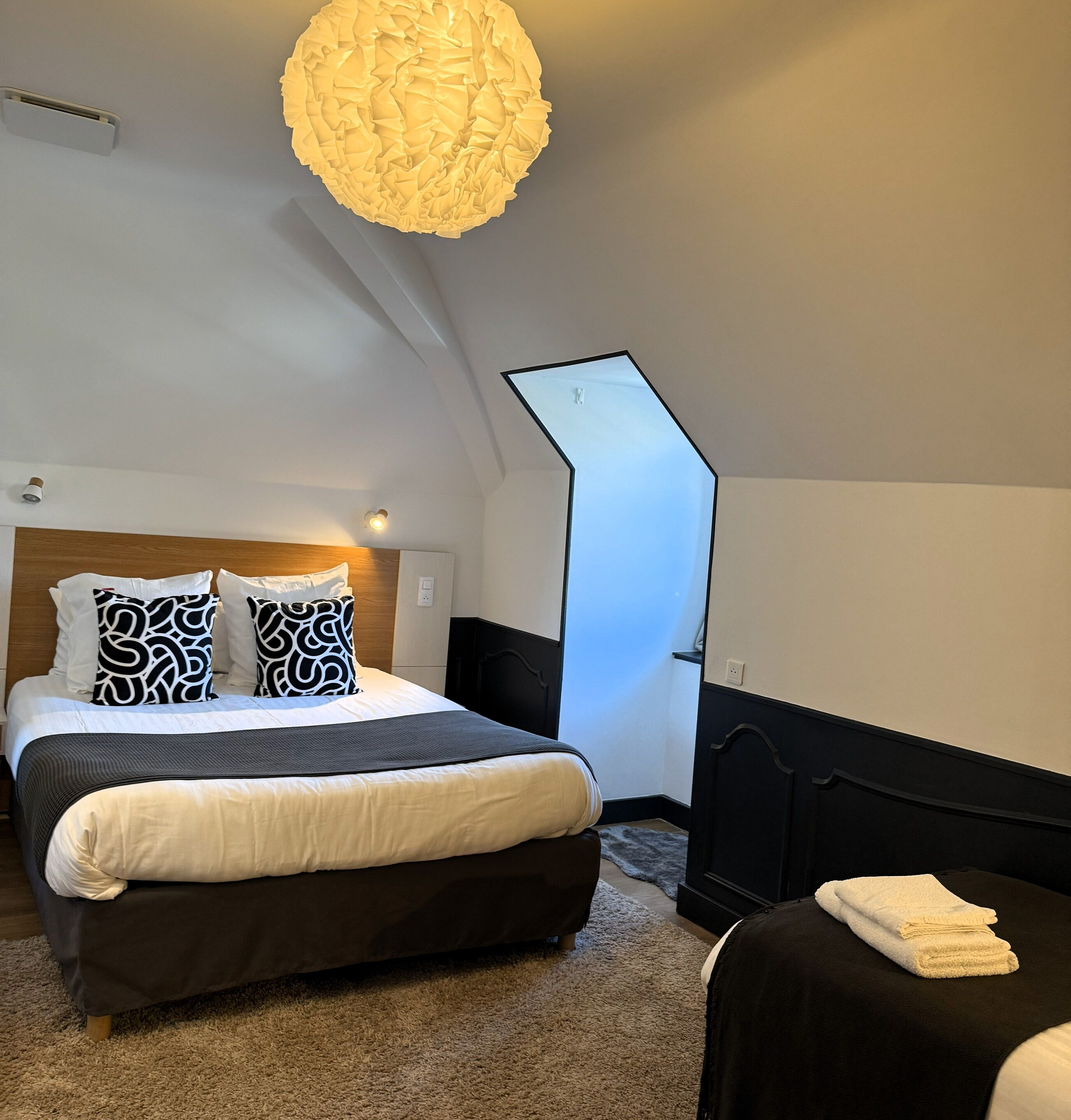 A modern bedroom with a large bed, two patterned pillows, a dark headboard, a unique light fixture, and a view of a staircase.