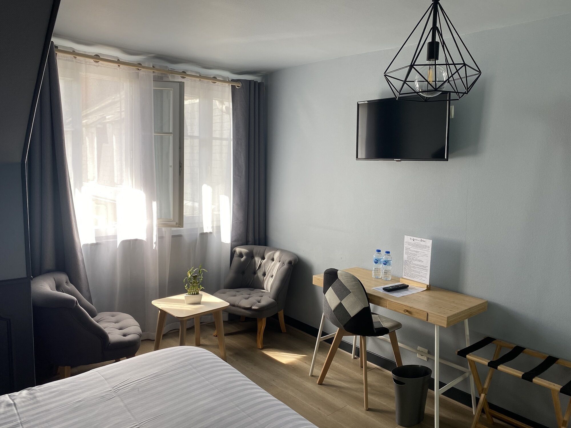 Une chambre d’hôtel comprenant un lit, deux fauteuils, une petite table avec une plante, un bureau avec une chaise, une télévision et une bouteille d’eau sur le bureau.