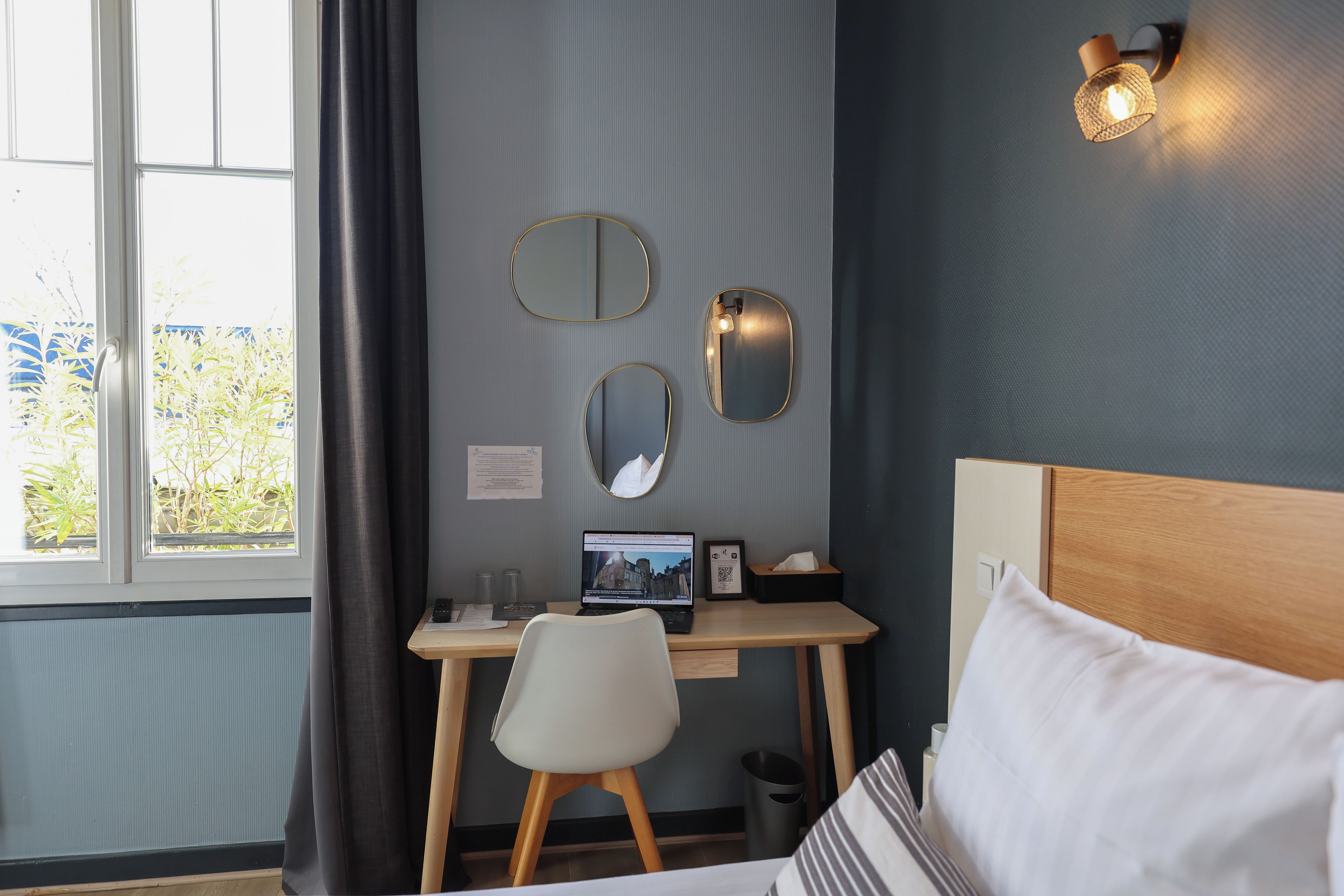 A hotel room with a bed, a desk with a laptop, two wall mirrors, a window with curtains, and a small trash bin.