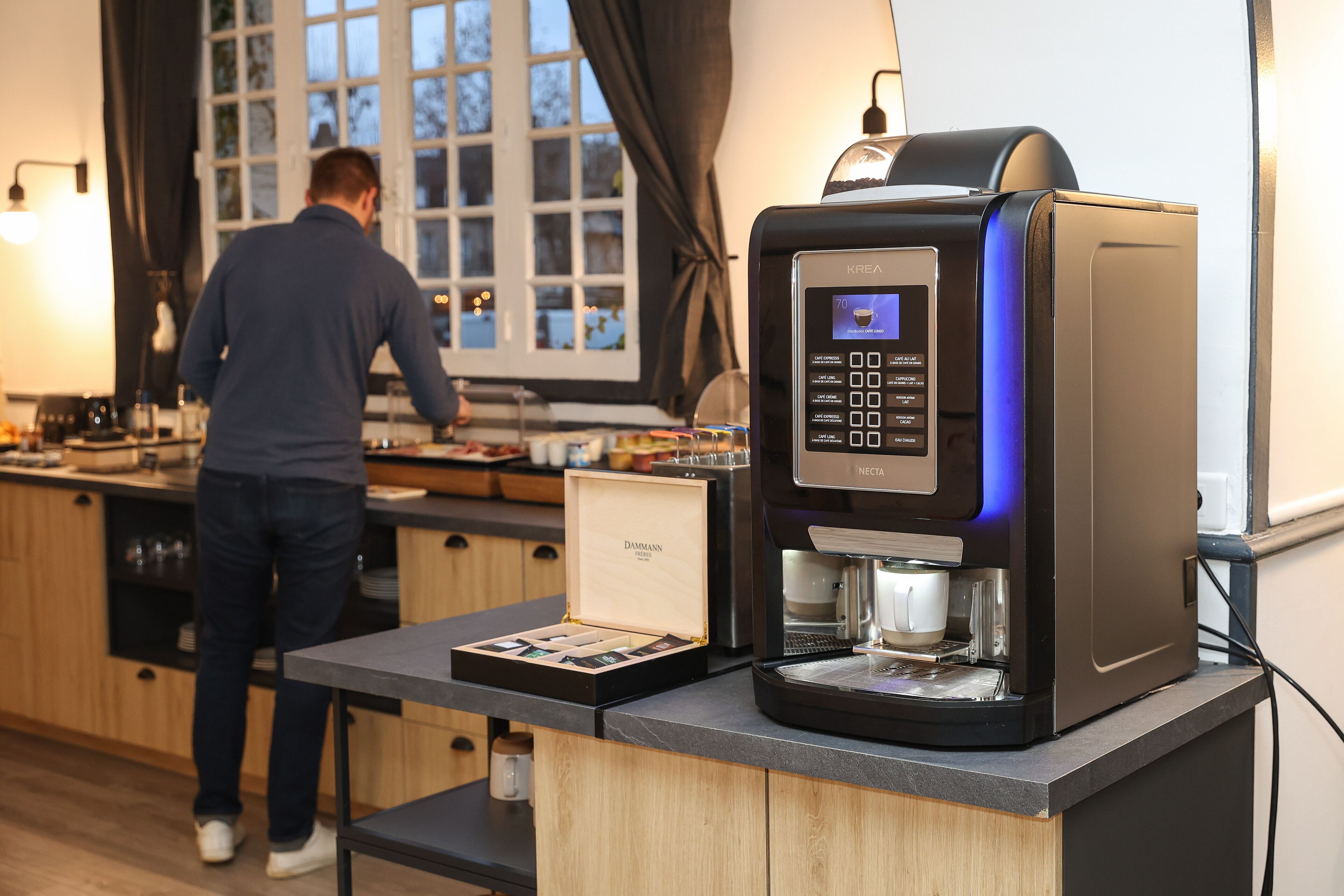 A coffee machine with a digital interface, a person using it, and a box of coffee pods on a counter.