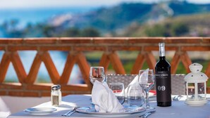 Mesa posta para dois com uma garrafa de vinho, copos e saleiro em terraço com vista para as colinas.