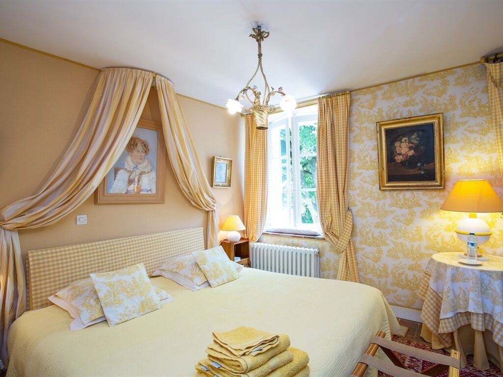 A bedroom with a bed, curtains, a chandelier, a window, a radiator, a table with a lamp, and a framed picture on the wall.