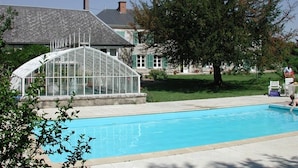 A swimming pool with a greenhouse structure, a house with a sloped roof, and a large tree in the background. - Château de la Rue (Suevres)
