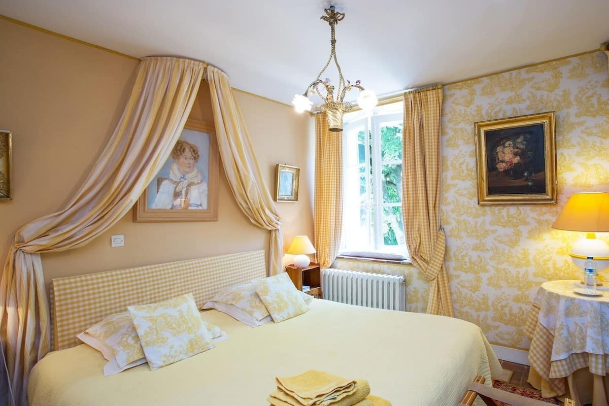 Une chambre à coucher avec un lit, un lustre, une fenêtre avec des rideaux, un radiateur et une table avec une lampe.