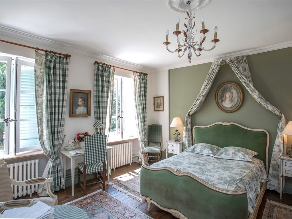 A bedroom with a green canopy bed, a chandelier, checkered curtains, and a view of the outdoors.