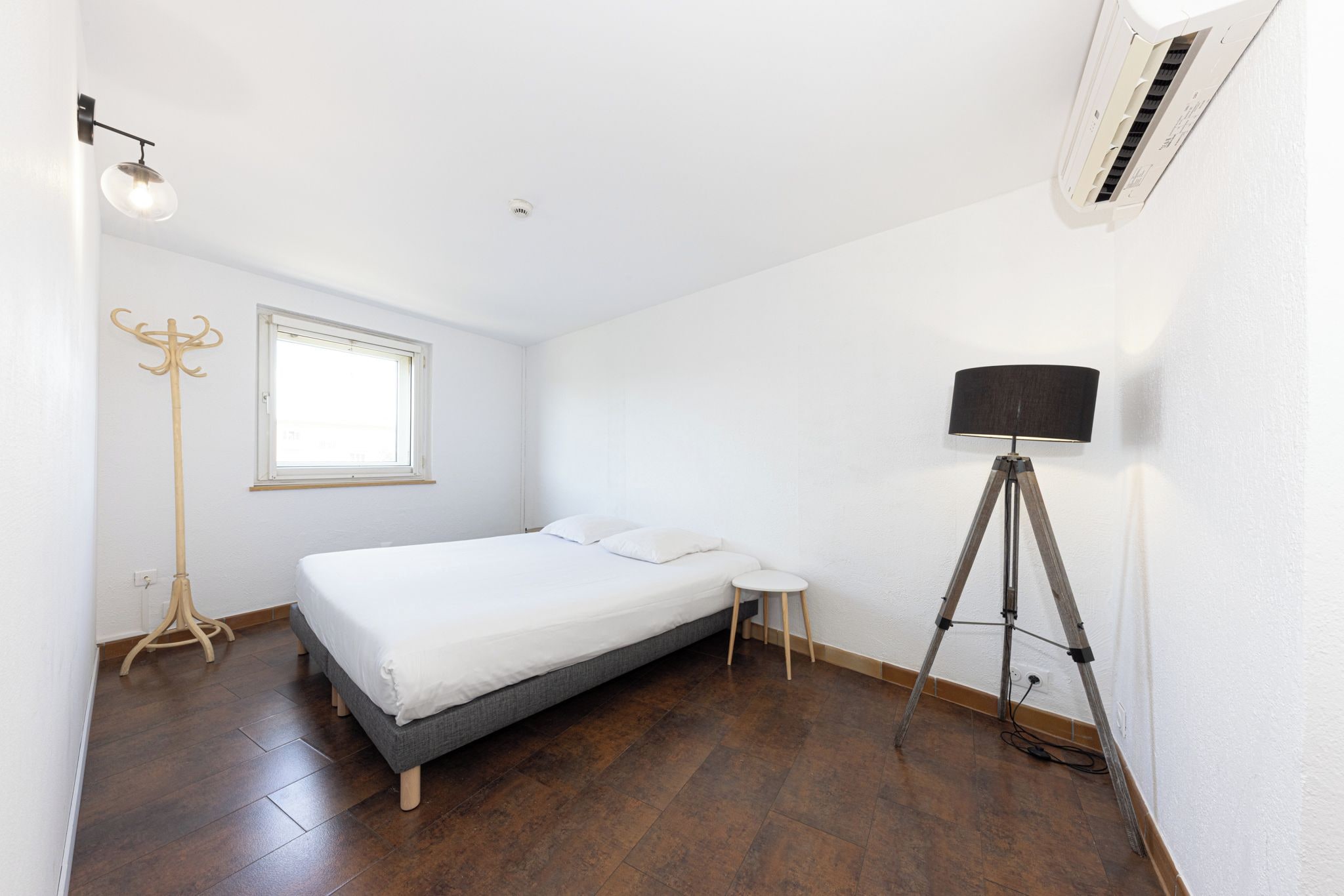 A bedroom with a bed, a window, a floor lamp, and an air conditioning unit.