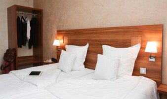 Habitación de hotel con cabecera de madera, ropa de cama blanca, mesita de noche con lámpara y perchero con ropa colgada.