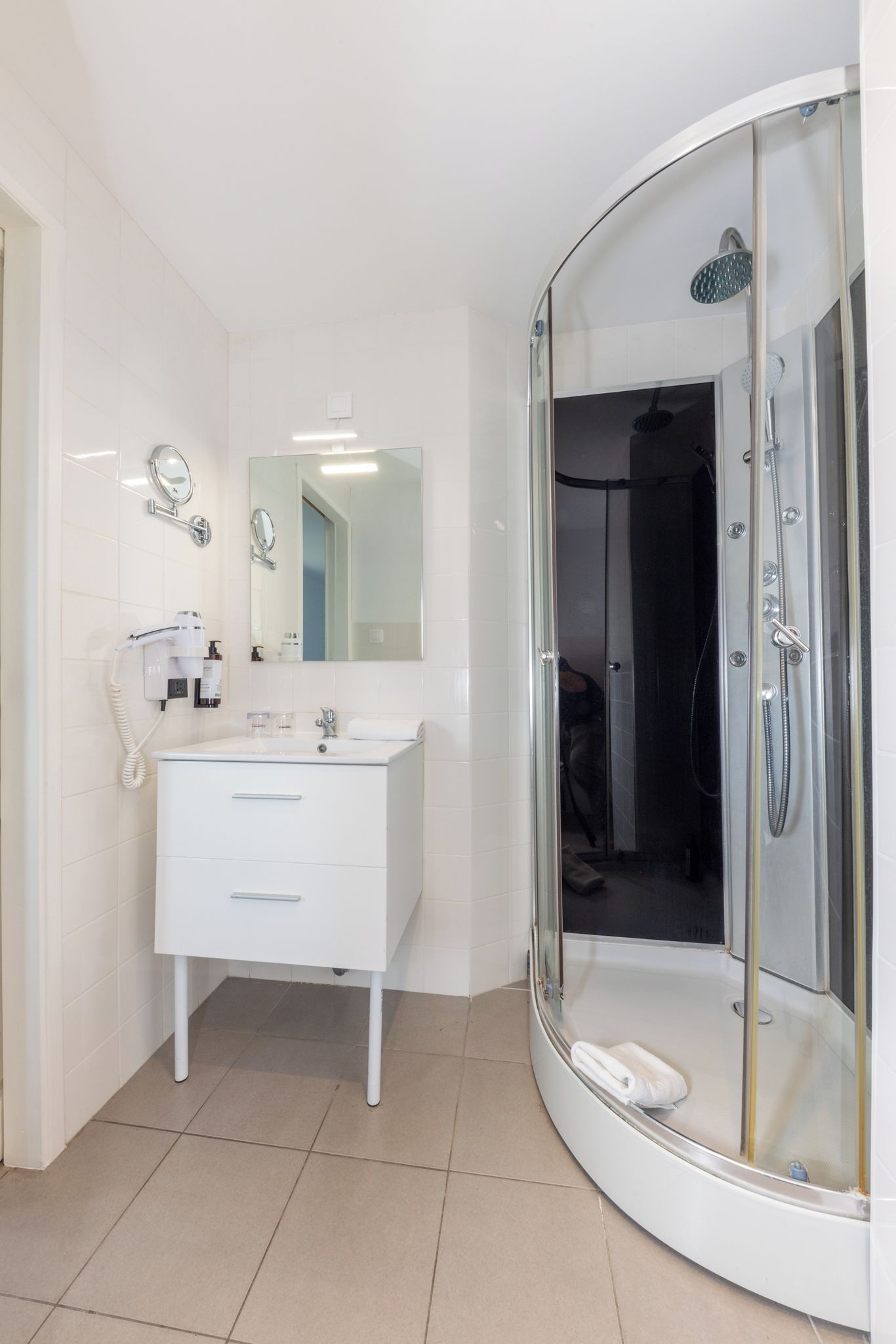 Une salle de bain moderne avec un meuble-lavabo blanc, un grand miroir, une douche à parois de verre et un sèche-cheveux fixé au mur.