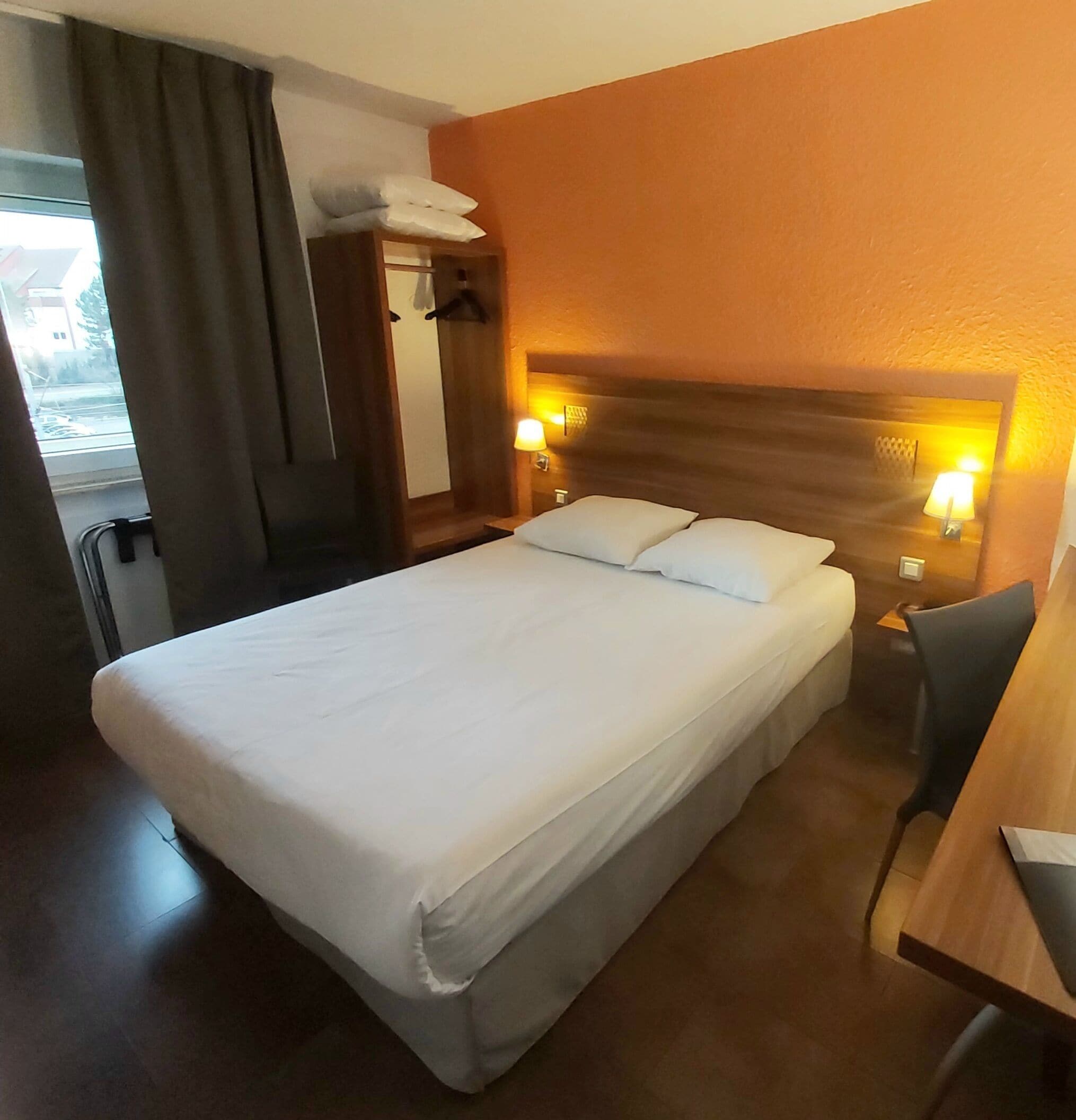 A hotel room with a large bed, two bedside lamps, a wooden headboard, a desk with a chair, and a window with curtains.