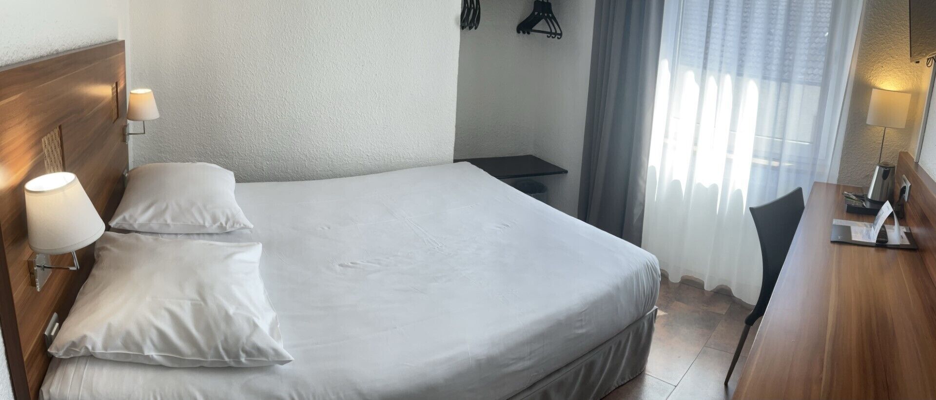 A hotel room with a large bed, two bedside lamps, a small table, and a window with curtains.