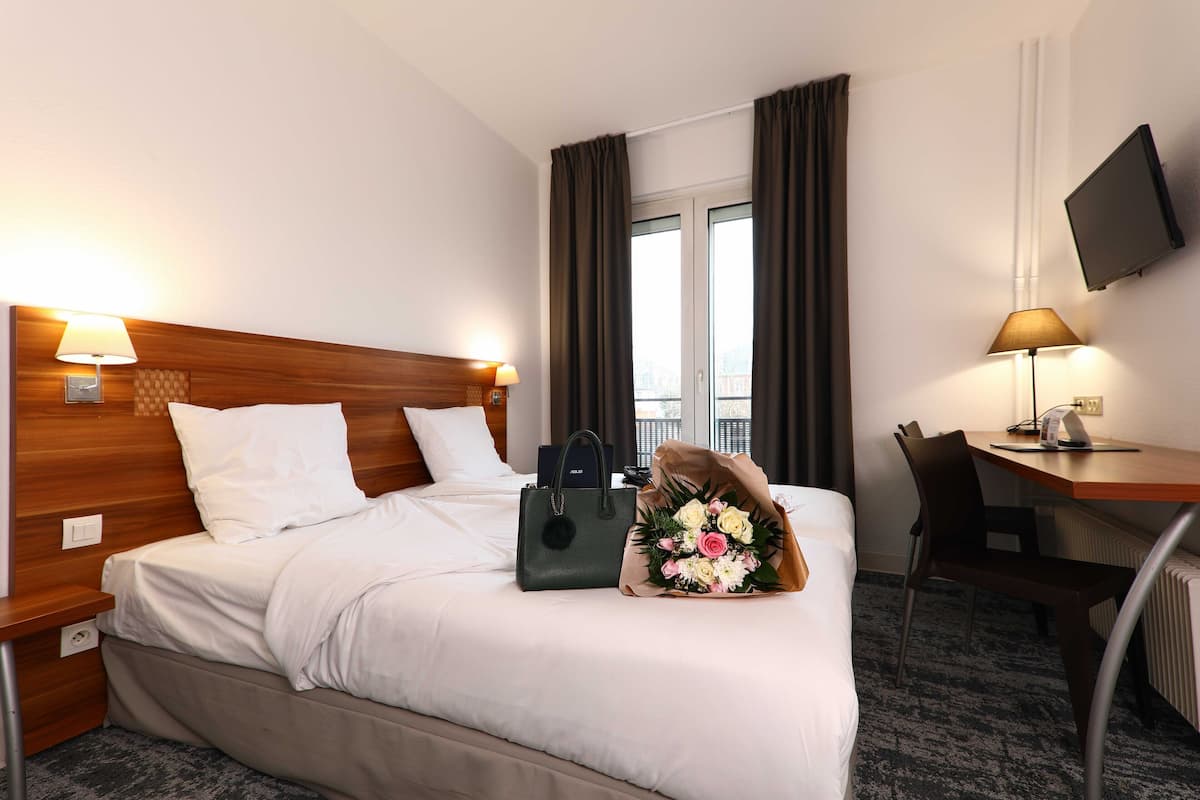 Habitación de hotel con cama, escritorio, silla, televisor y ventana con cortinas.