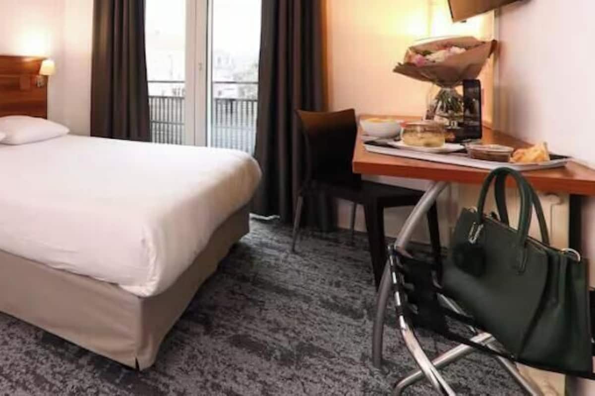 Una habitación de hotel con cama, escritorio, silla, lámpara y bolso.