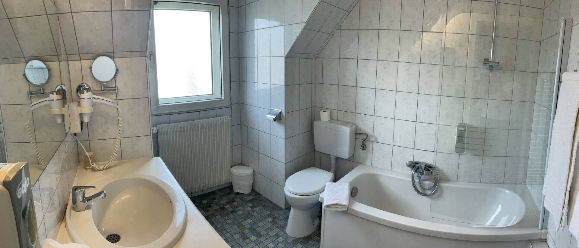 A modern bathroom with a bathtub, toilet, and sink, featuring a window and tiled walls.