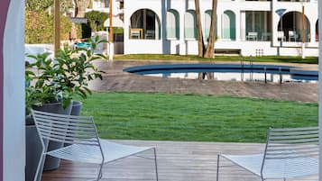 Vista a través de un arco de un patio con piscina, un edificio blanco con varios balcones y un césped verde.
