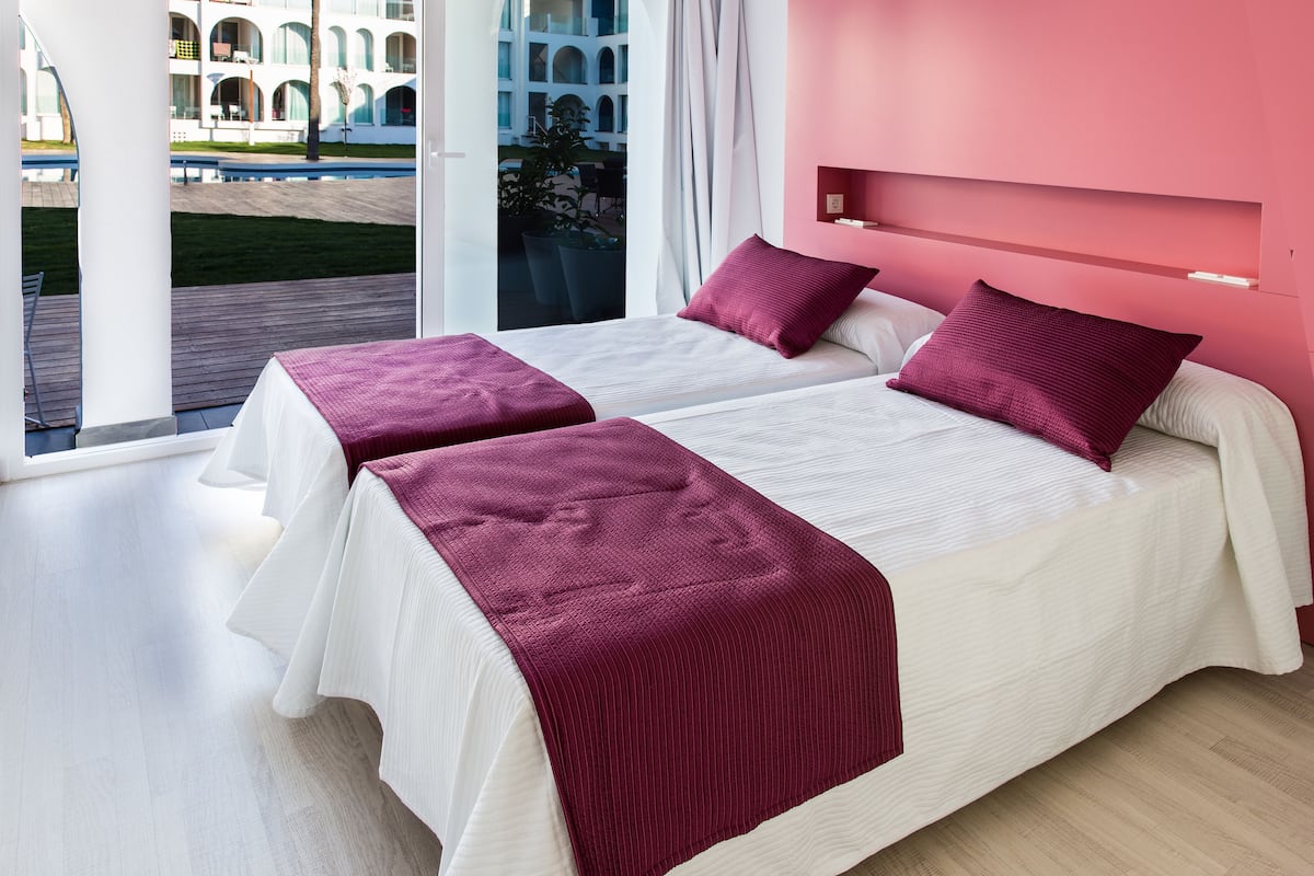 A modern hotel room with two beds, a large window, and a view of a building with arches.