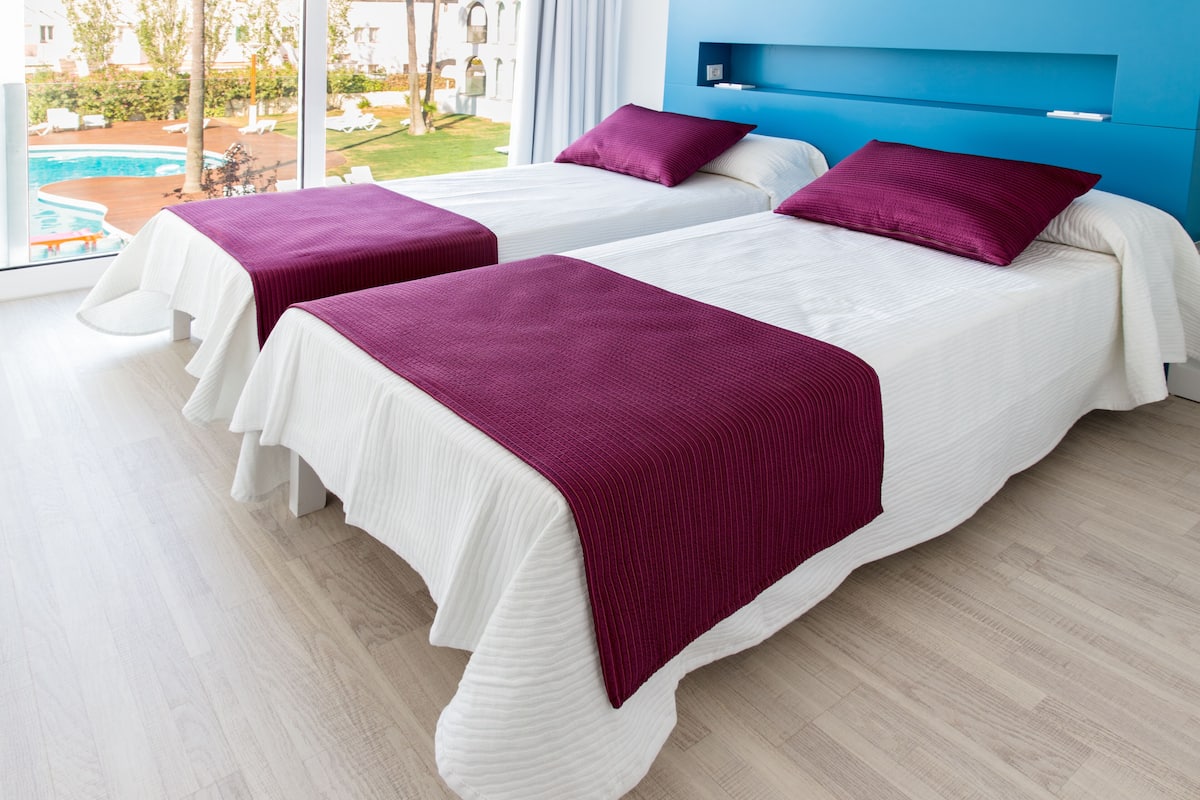 Two beds with white bedding and purple throw blankets, a blue headboard, and a view of a pool and greenery outside.