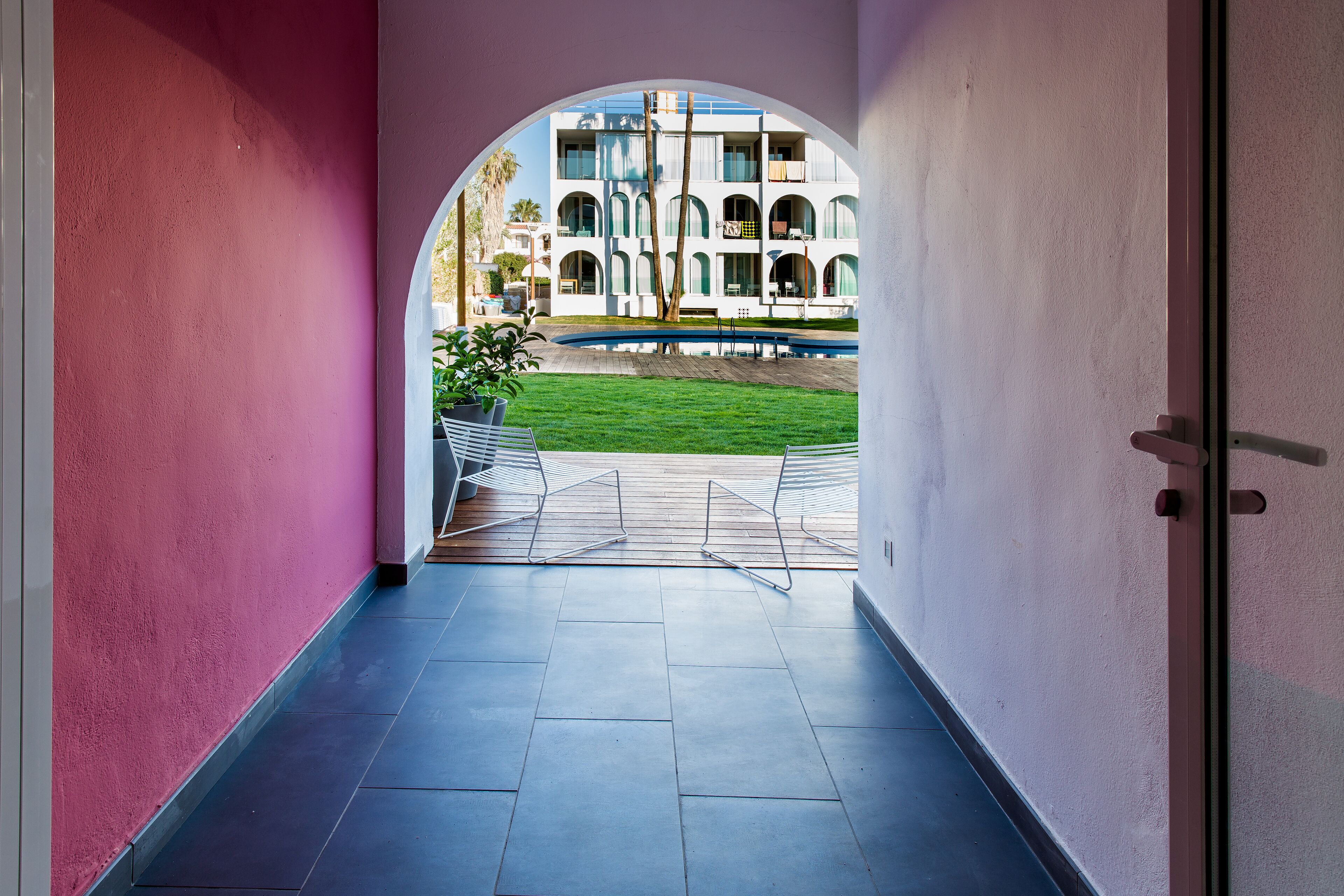 A corridor with a tiled floor leading to an outdoor area with a pool and a building with arched windows in the background.