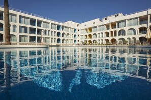 Uma piscina grande com água azul cristalina, rodeada por um edifício de vários andares com varandas e janelas em arco.