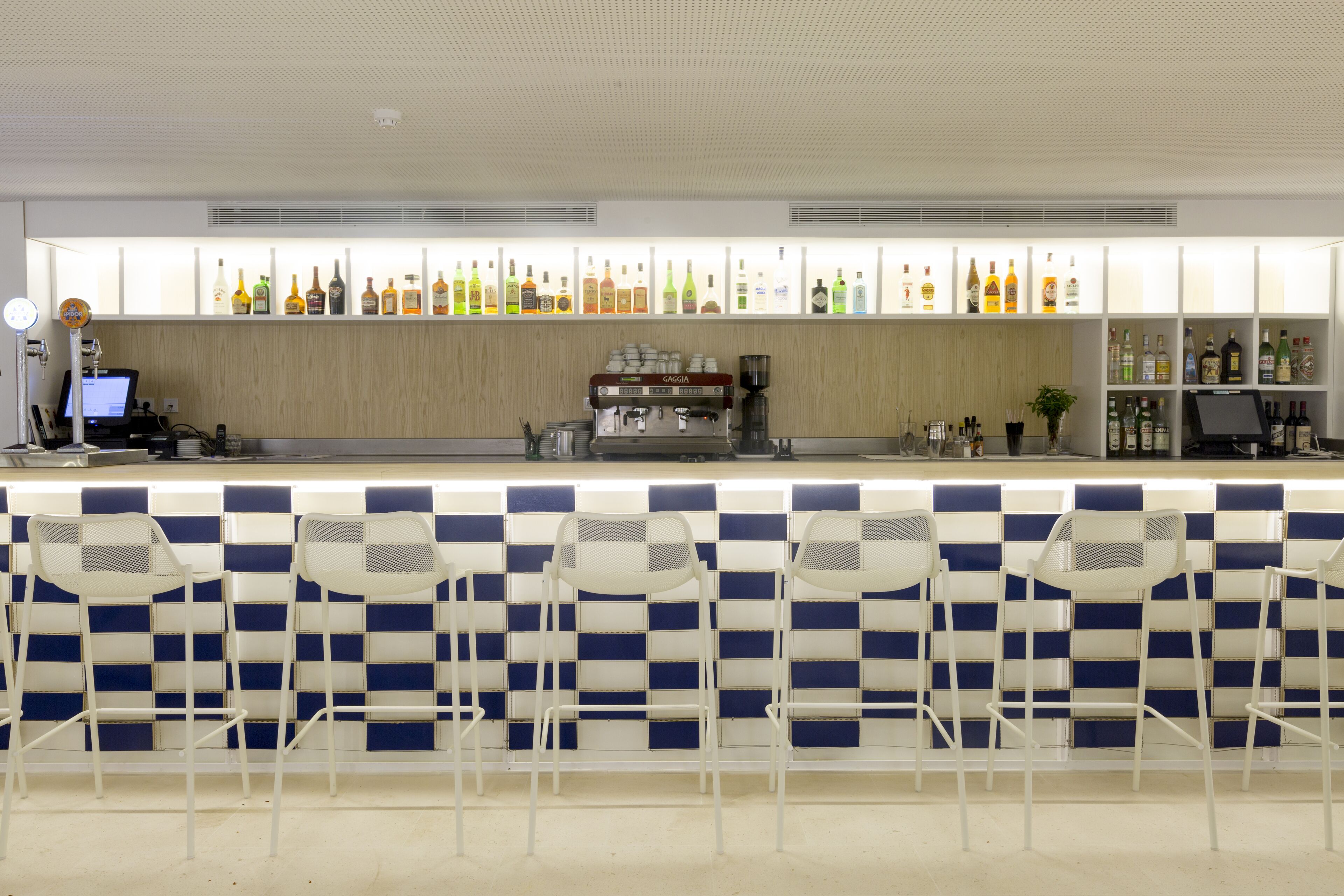 Een bar met een blauw-wit geruit wandpaneel, een koffieapparaat en diverse flessen op planken.