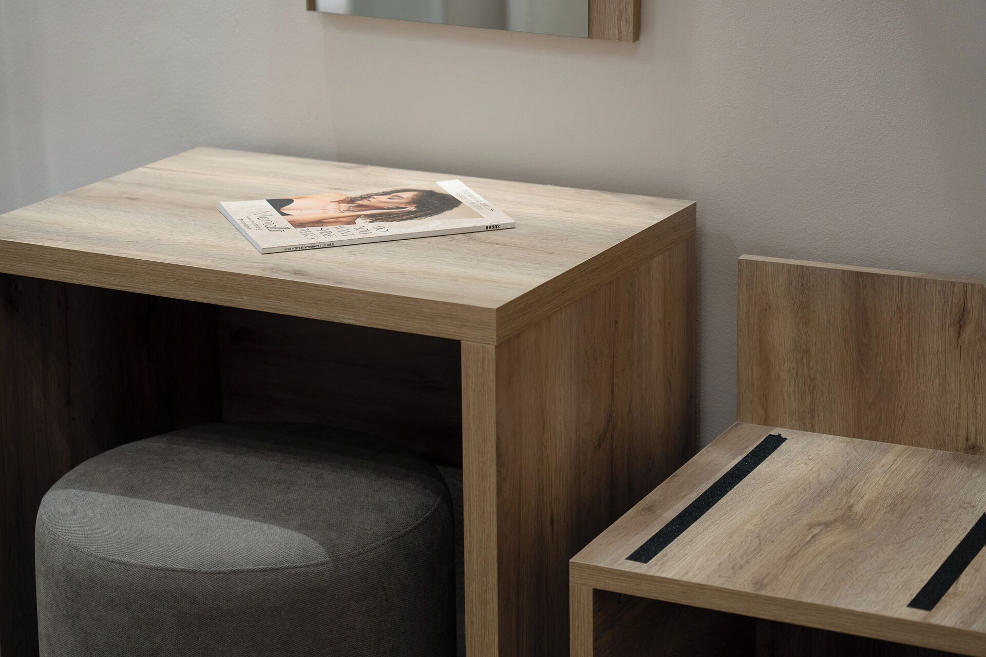 A small wooden table with a magazine on it, a grey upholstered stool, and a wooden chair with a black stripe.