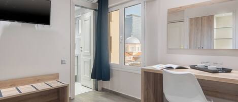 Habitación moderna con televisión de pantalla plana, un banco de madera, un escritorio con una silla, un ventanal con vista a un edificio y un espejo sobre el mueble del espejo.