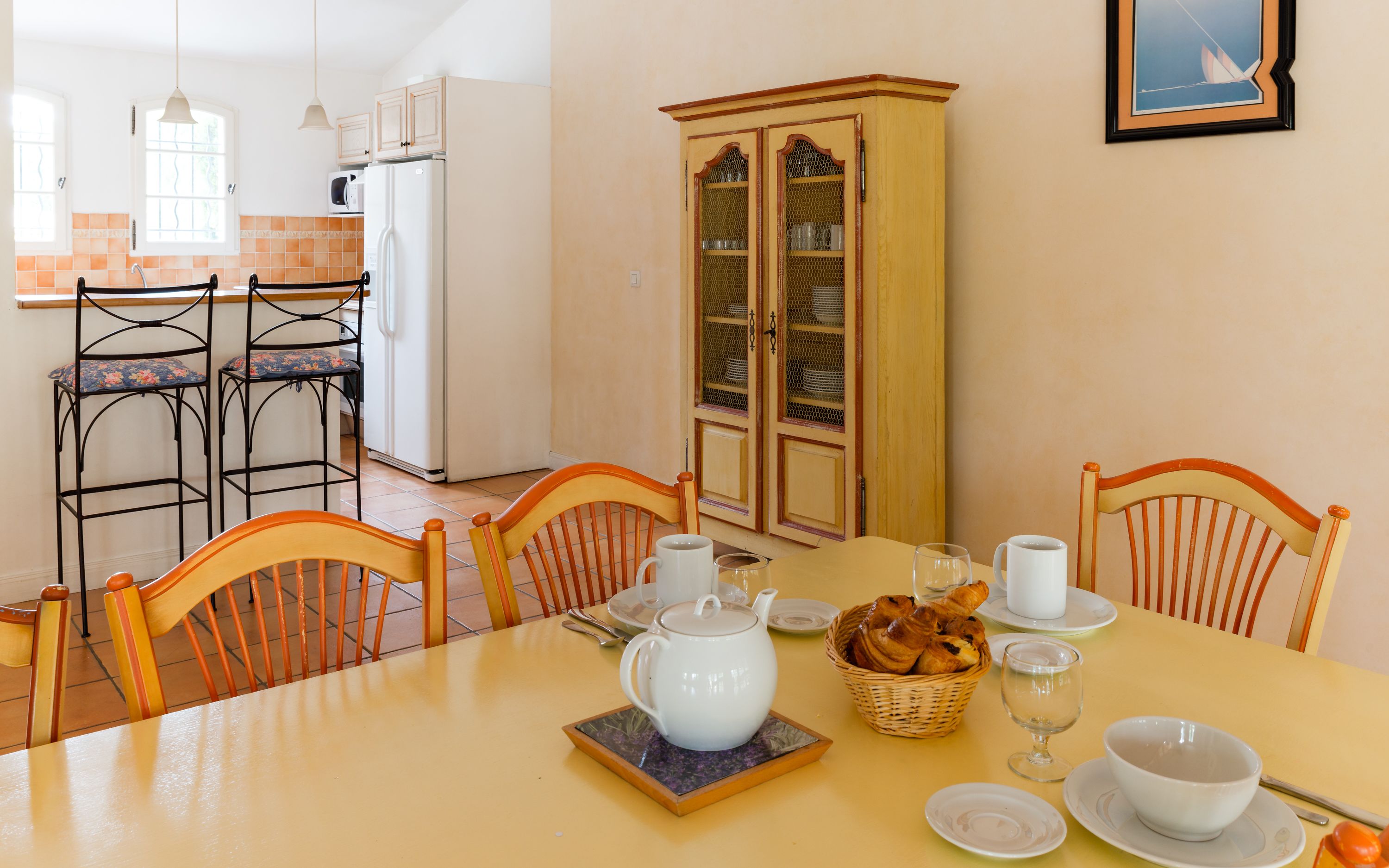 A dining area with a table set for four, chairs, a cabinet with glass doors, and a kitchen in the background.
