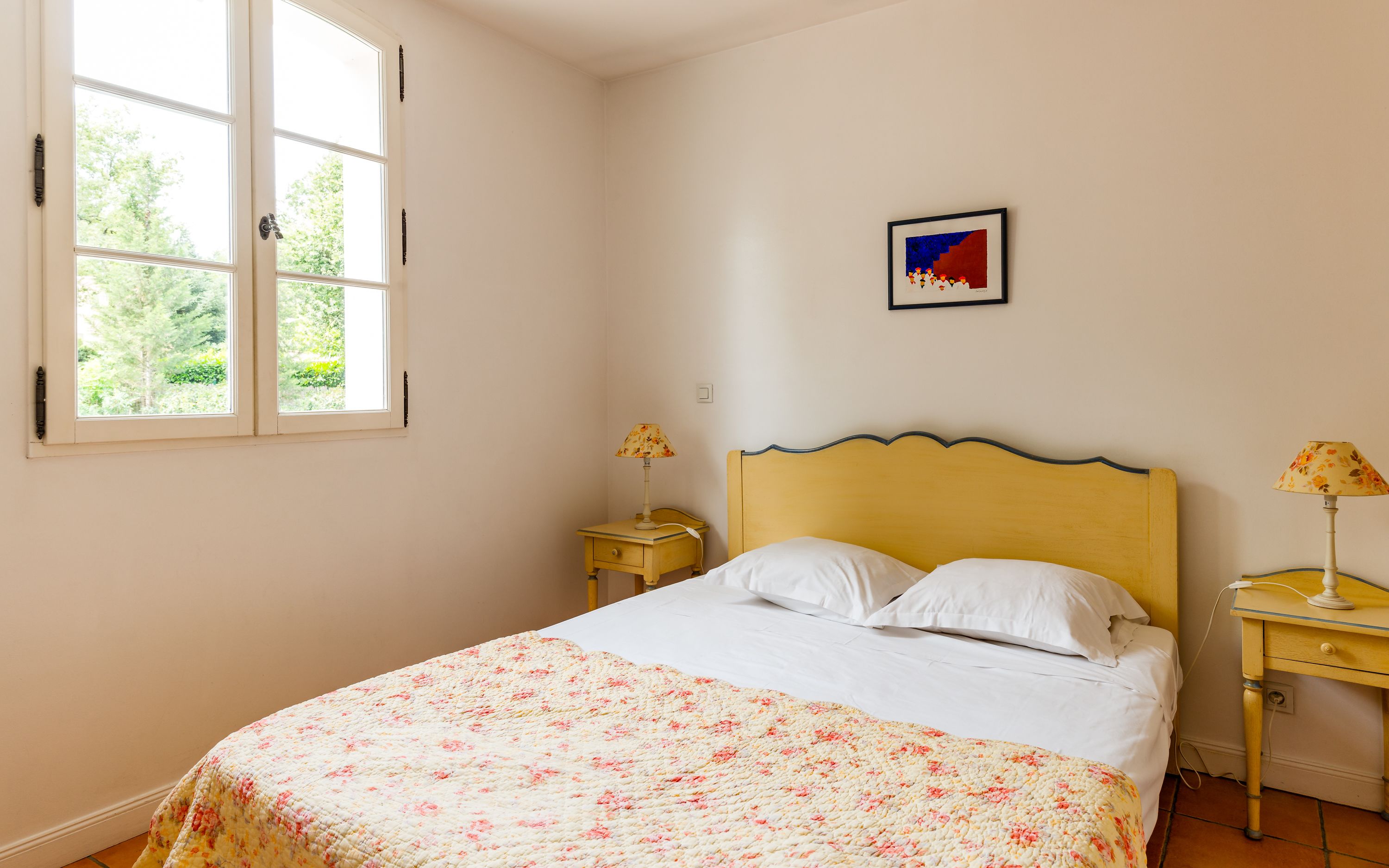 A bedroom with a bed, two bedside tables, a window, and a wall-mounted picture.