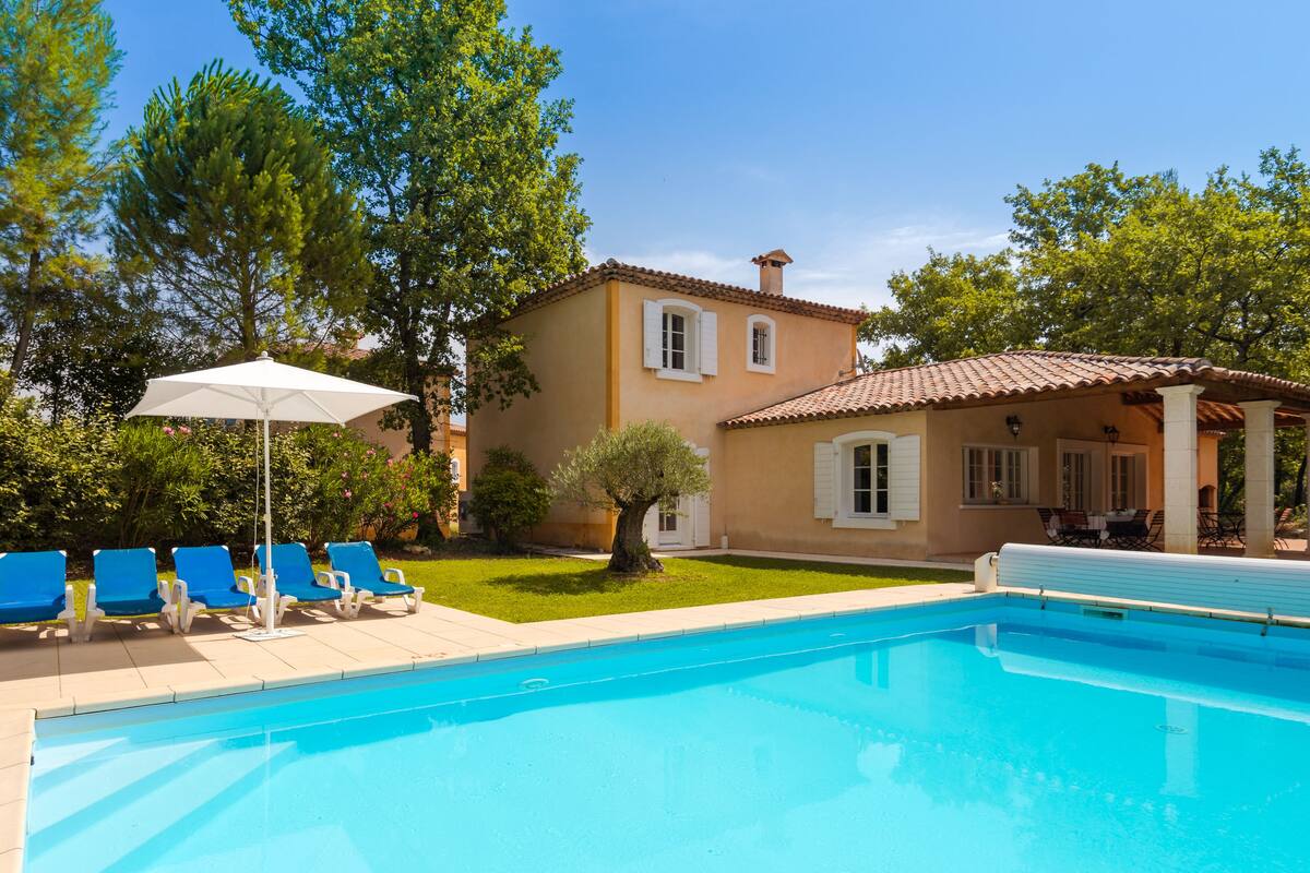 Ein Schwimmbecken mit blauem Wasser, umgeben von einer Terrasse mit blauen Liegestühlen und einem weißen Sonnenschirm. Im Hintergrund ein Gebäude mit einem Ziegeldach sowie ein Garten mit Bäumen und Sträuchern.