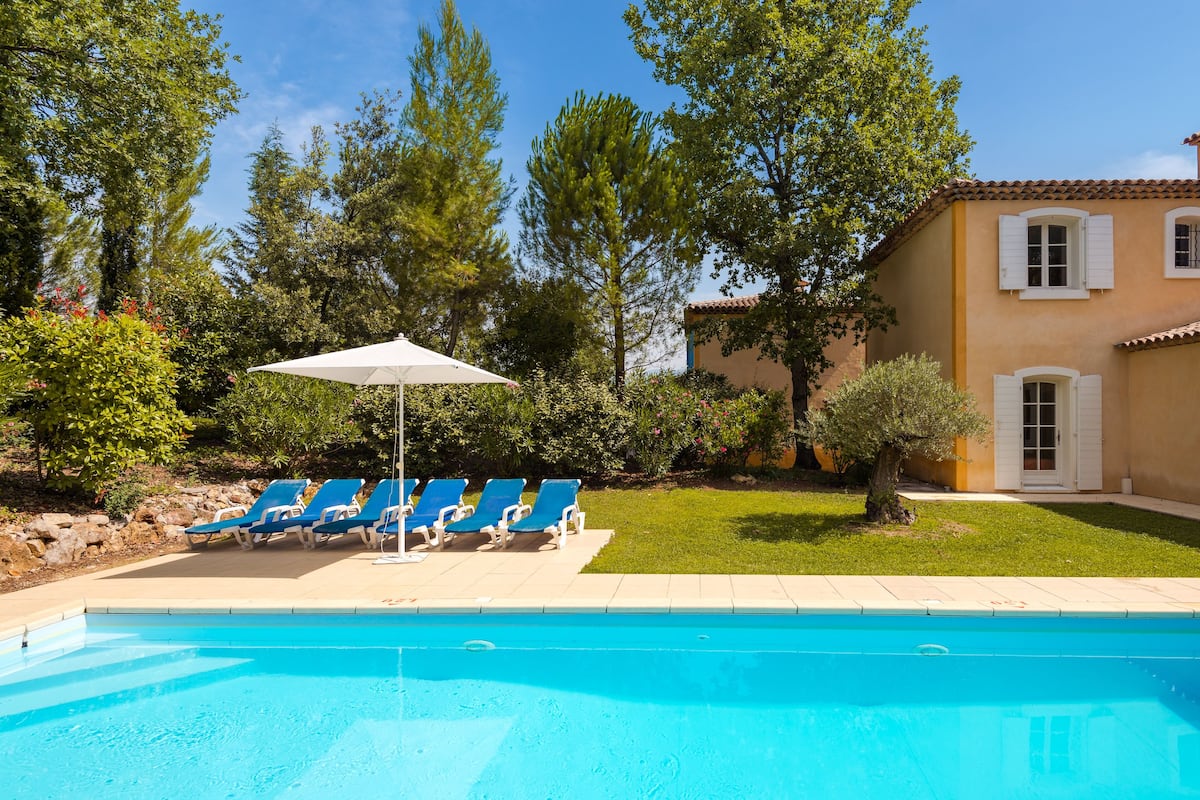 Ein Schwimmbecken mit blauem Wasser, umgeben von einem Garten mit Bäumen und einem Gebäude mit einem Ziegeldach.