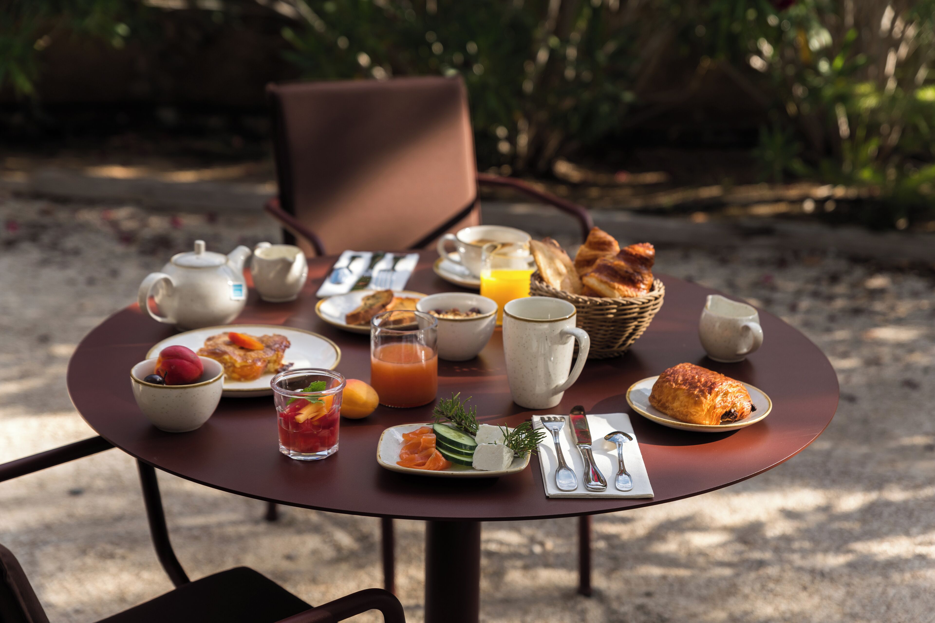 Una mesa redonda con alimentos para el desayuno, incluyendo cruasanes, una tetera, una taza y un vaso de zumo de naranja.
