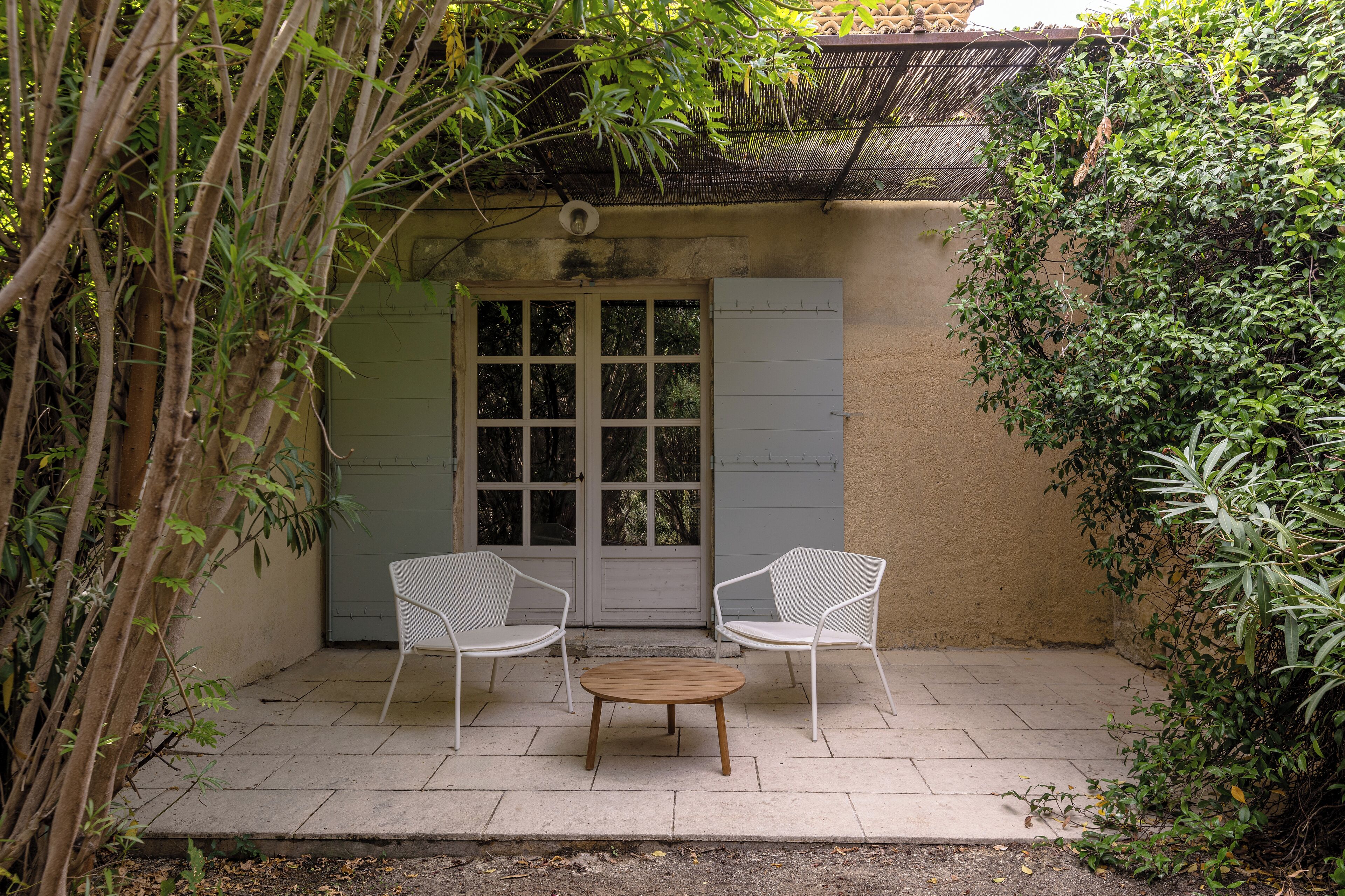 A patio with two white chairs and a round table, surrounded by greenery and a door with glass panels.