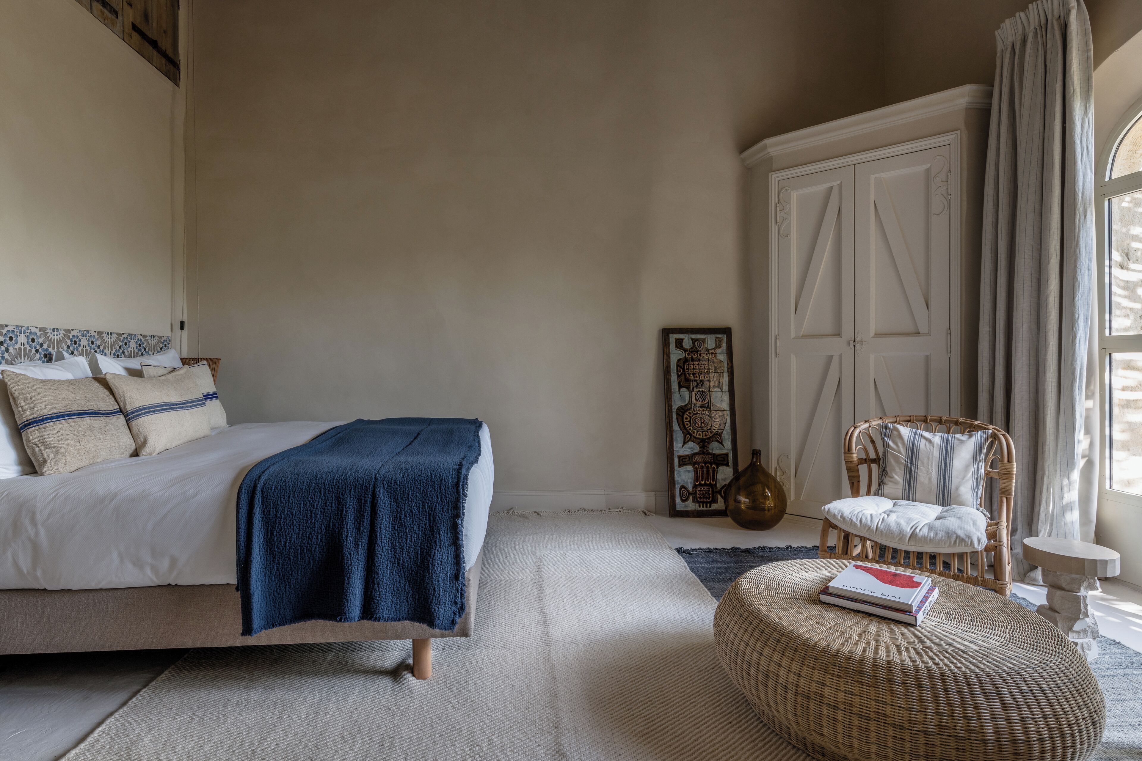 A bedroom with a bed, a wicker ottoman, a chair, a small table, and a wardrobe.