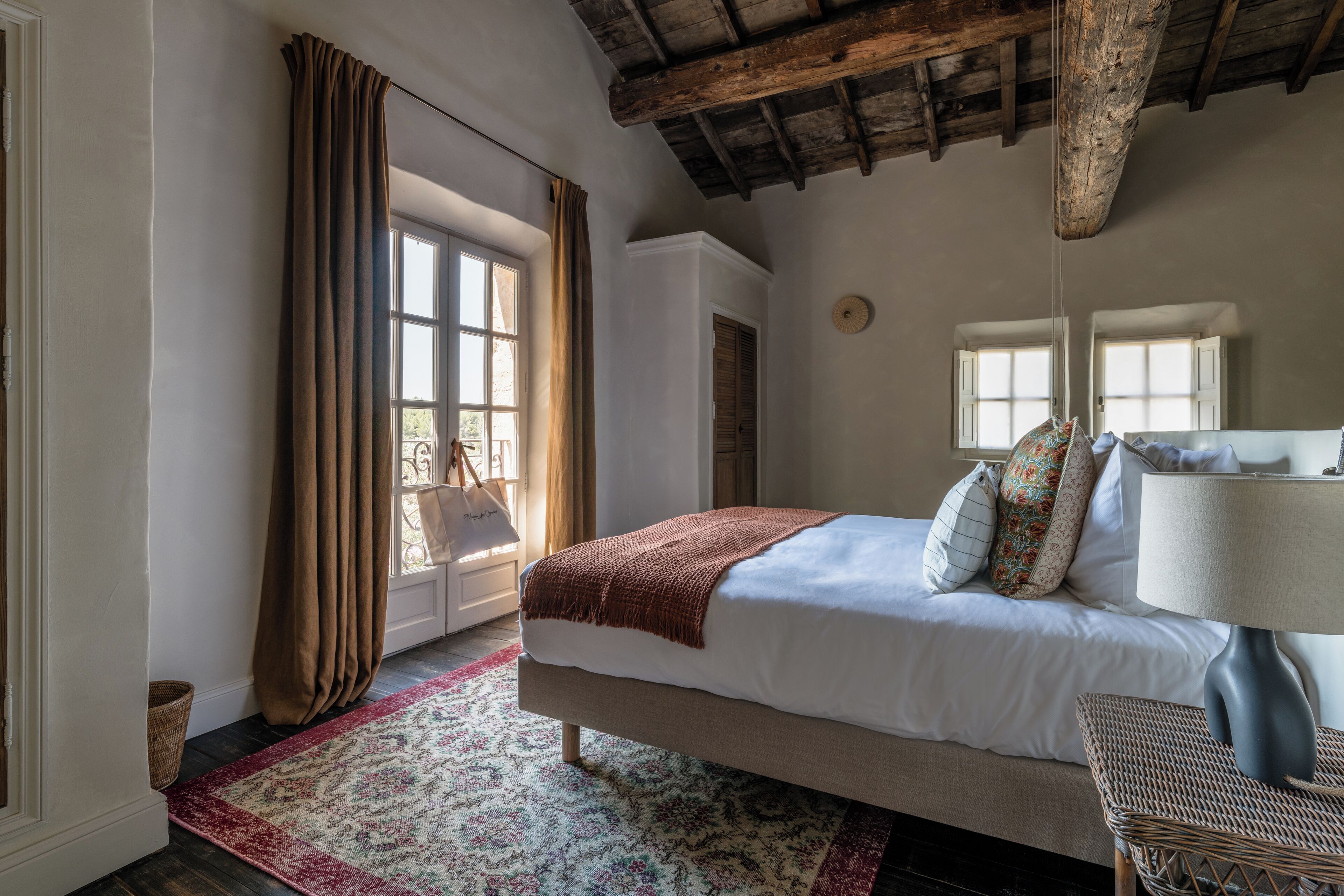Ein Schlafzimmer mit Bett, Lampe, Korbsessel und Fenster mit Vorhängen.
