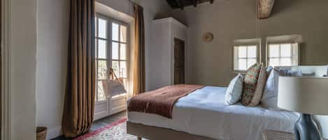 Ein Schlafzimmer mit Bett, Lampe, Korbsessel und Fenster mit Vorhängen.