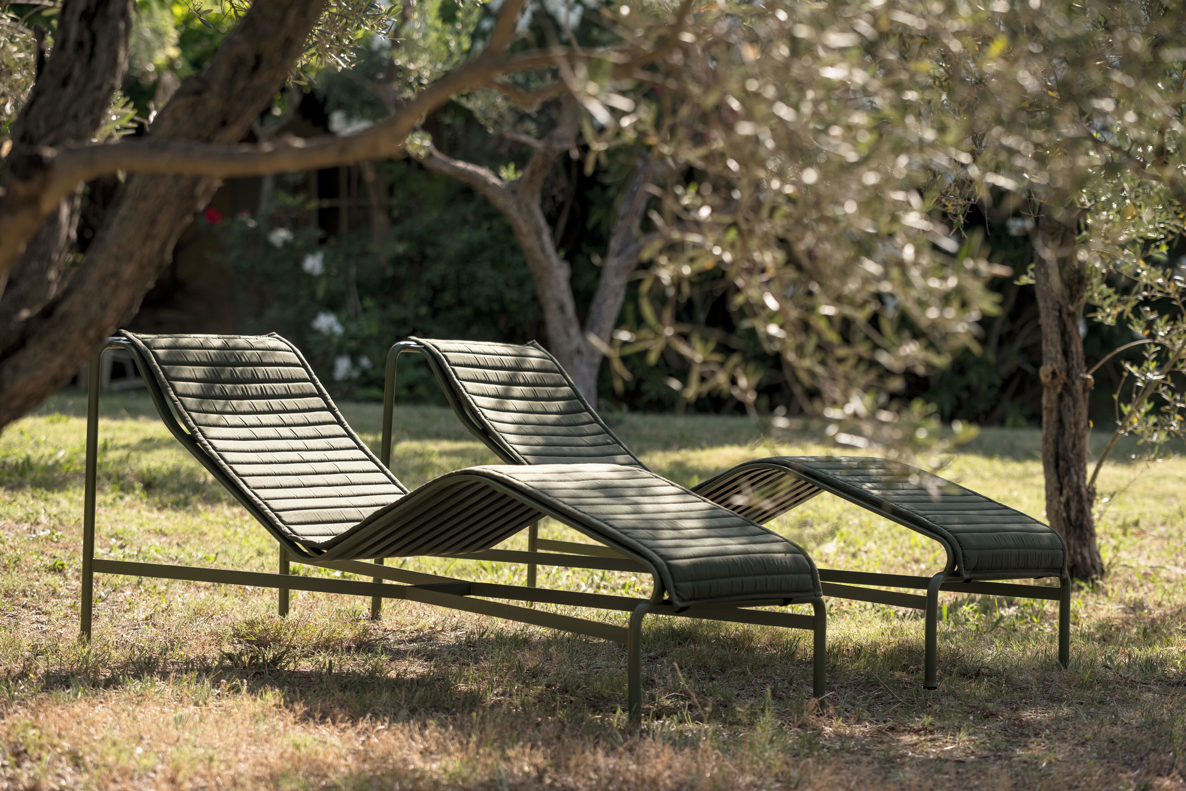 Two outdoor lounge chairs with metal frames and cushioned seats, positioned on a grassy area with trees in the background.