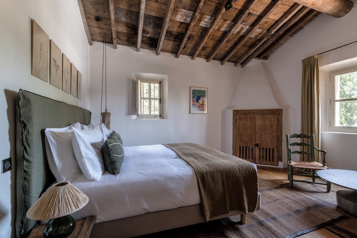 Un dormitorio con techo de madera, una cama con ropa blanca, una silla verde y una ventana con cortinas.