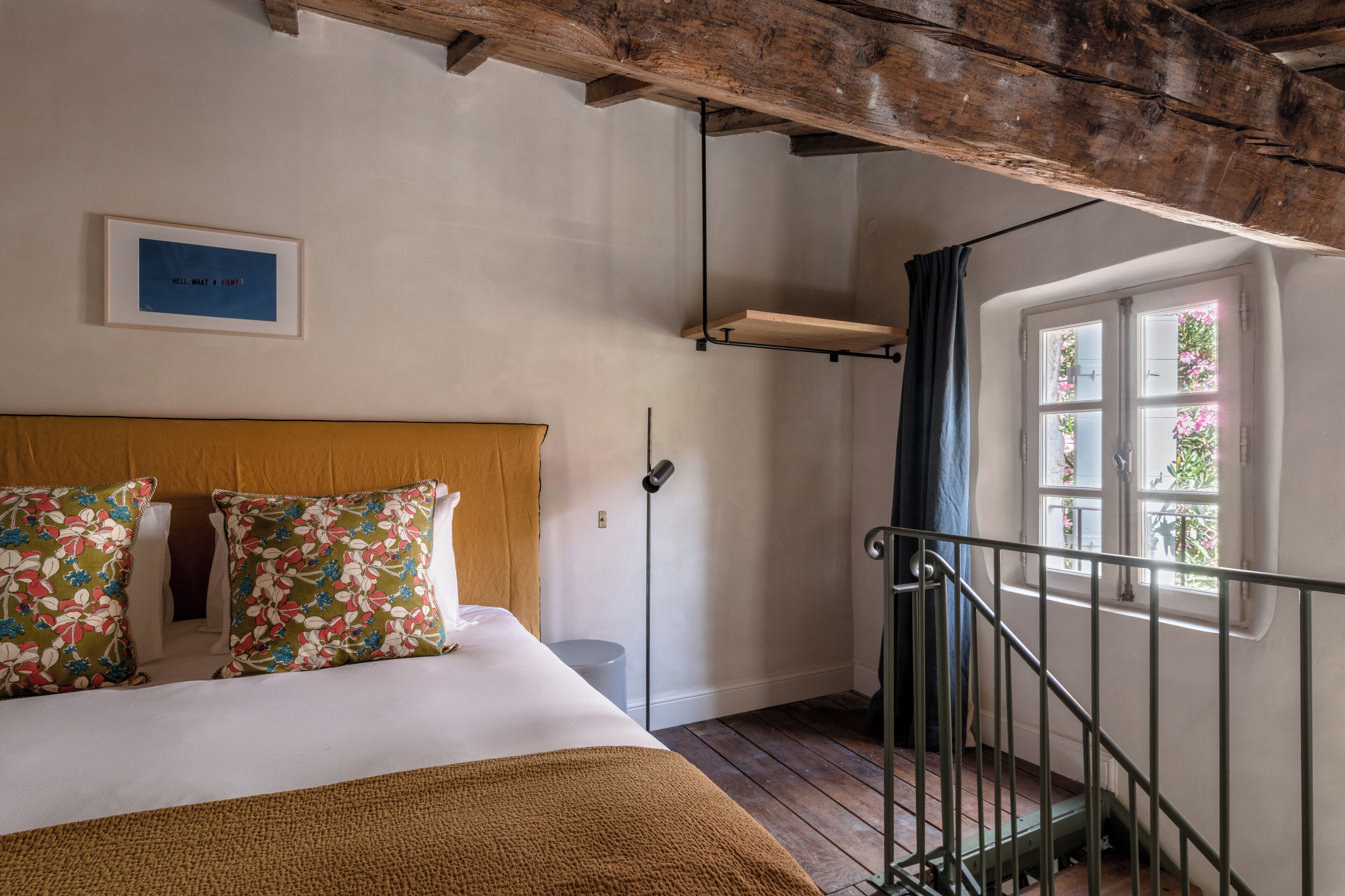 Un dormitorio con cabecera de madera, almohadas estampadas, un cuadro enmarcado, una ventana con cortinas y una escalera.