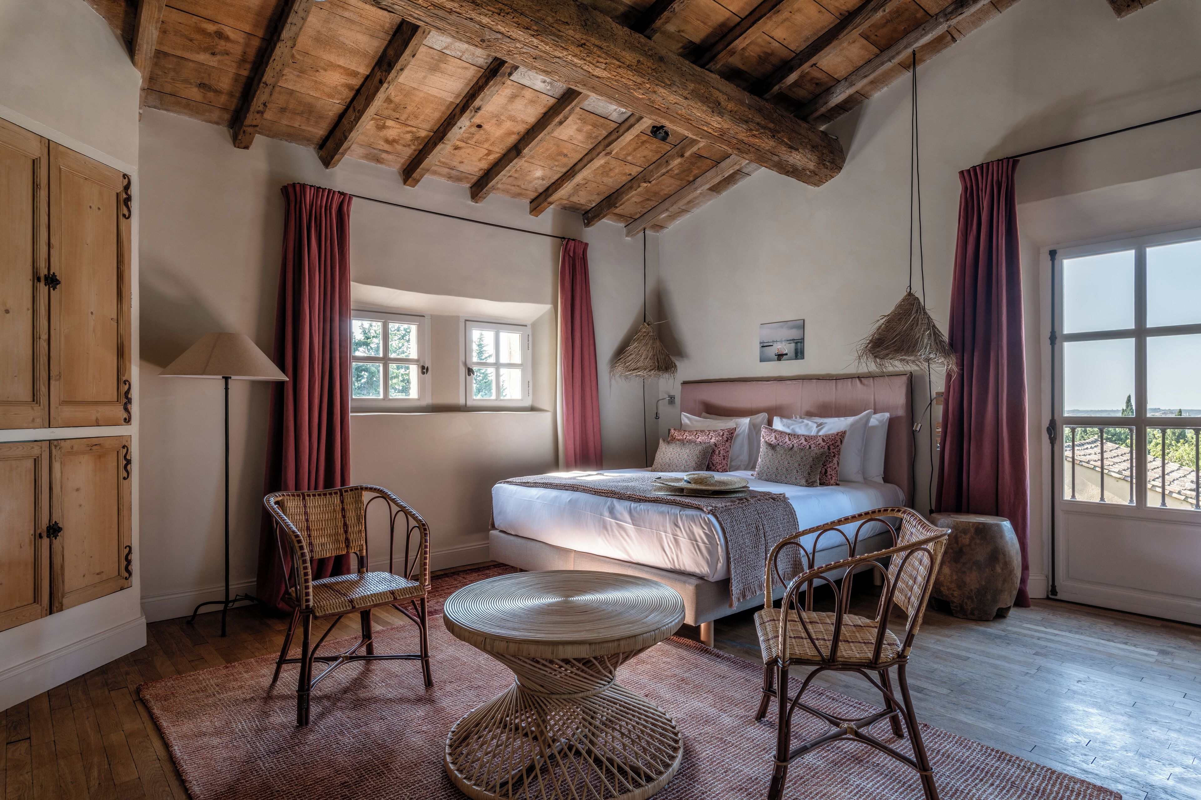 A bedroom with a wooden ceiling, a bed with pillows, a wicker chair, a round table, and a window with curtains.