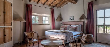 A bedroom with a wooden ceiling, a bed with pillows, a wicker chair, a round table, and a window with curtains.