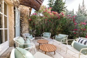 Un patio exterior con mobiliario de mimbre blanco, una pared de piedra y plantas florecientes de color rosa.