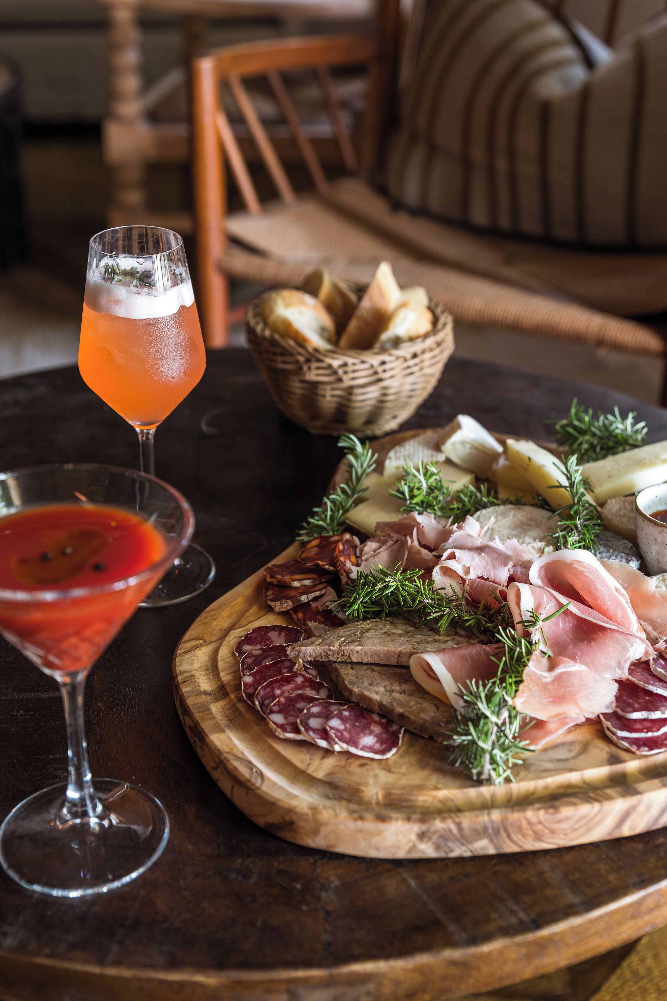 A wooden board with various meats and cheeses, a glass of red drink, and a basket of bread on a table.