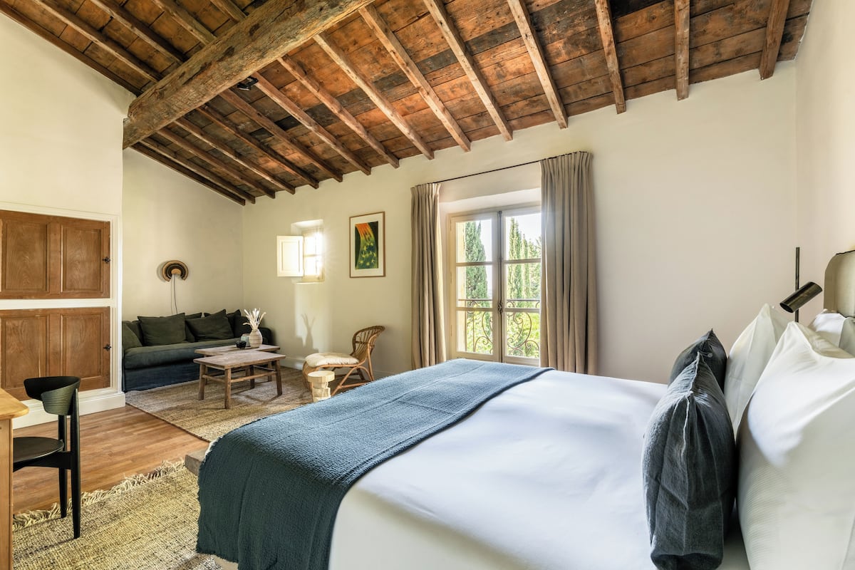 Un dormitorio con techo de madera, una cama con ropa de cama blanca, un escritorio de madera, una silla, un sofá y una ventana con cortinas.