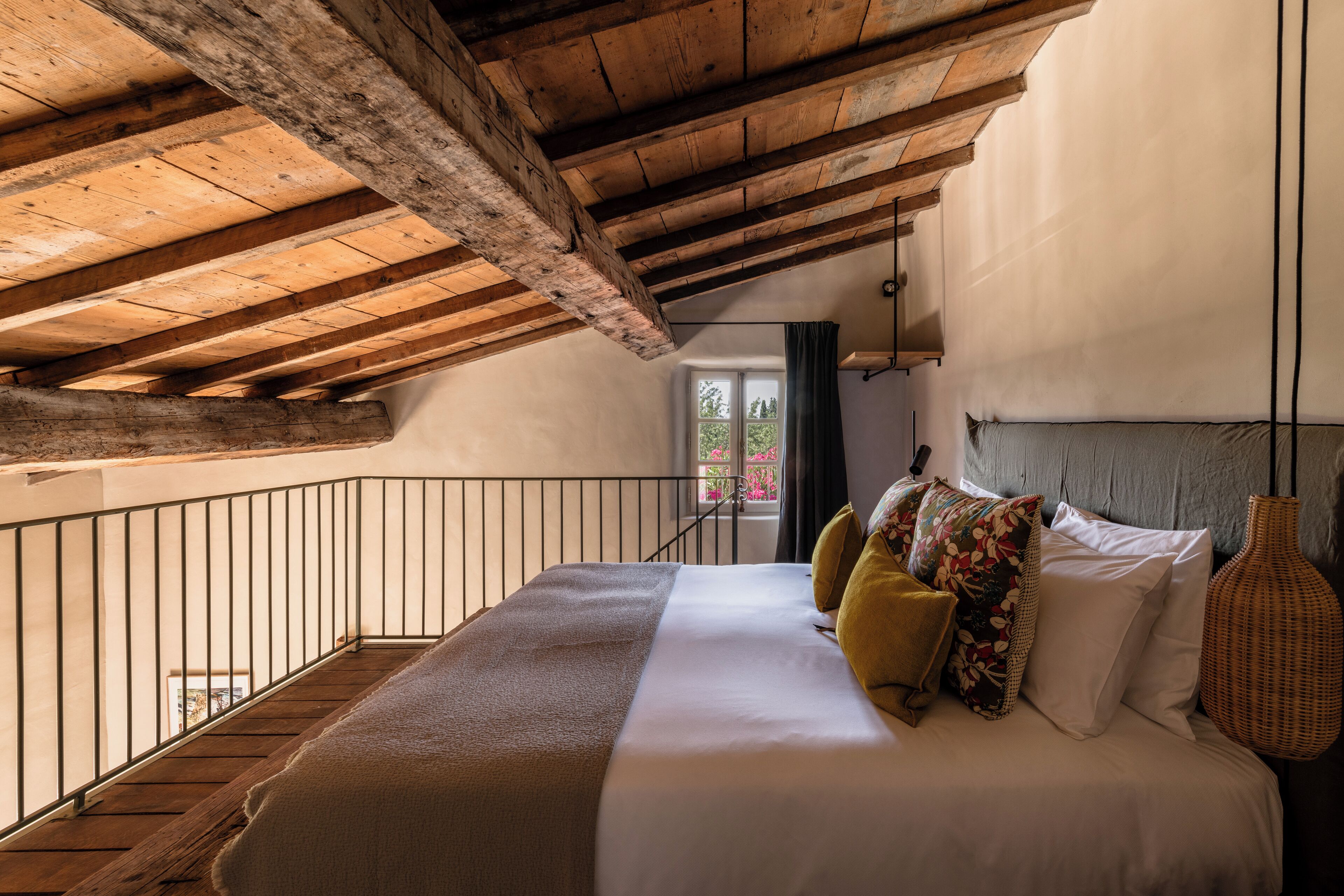 A bedroom with a wooden ceiling, a bed with multiple pillows, a vase, and a window with a view of greenery.